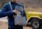 A man in a blue jacket is placing a power bank into a grey shoulder bag. A yellow car is parked in the background.