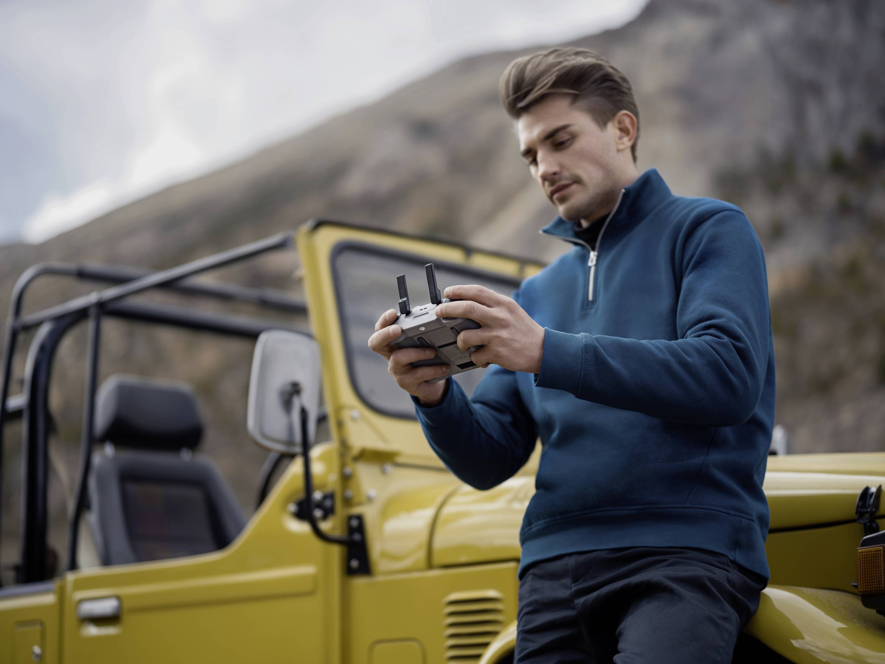 A man stands beside a yellow off-road vehicle and operates a drone with a controller against a mountainous backdrop.