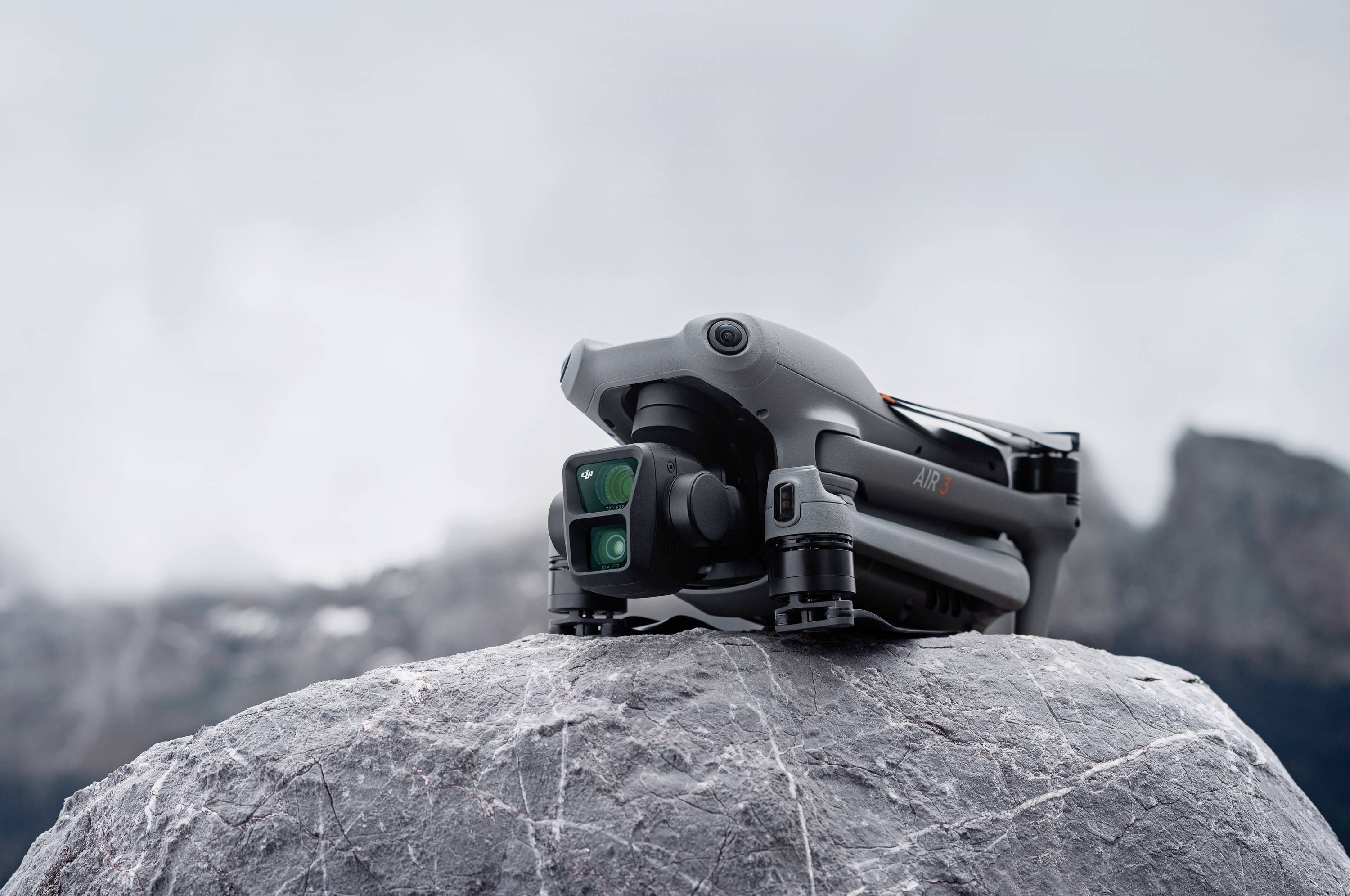 A grey drone rests on a rock, with a misty mountain landscape in the background.