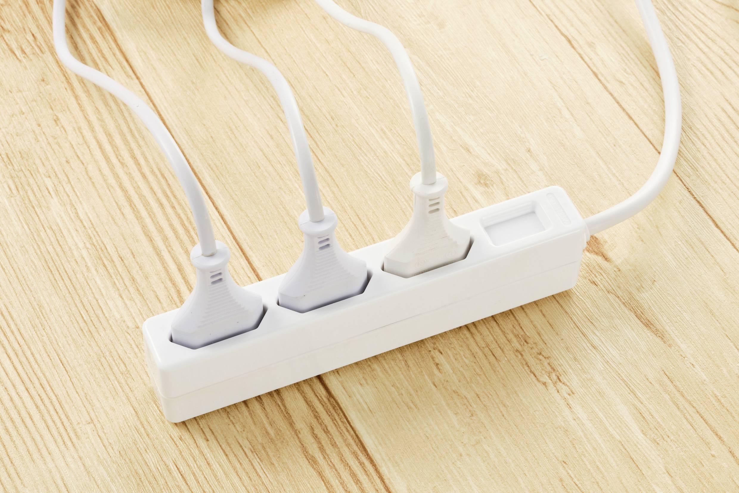 Three white plugs are plugged into a white power strip, which is lying on a wooden floor.