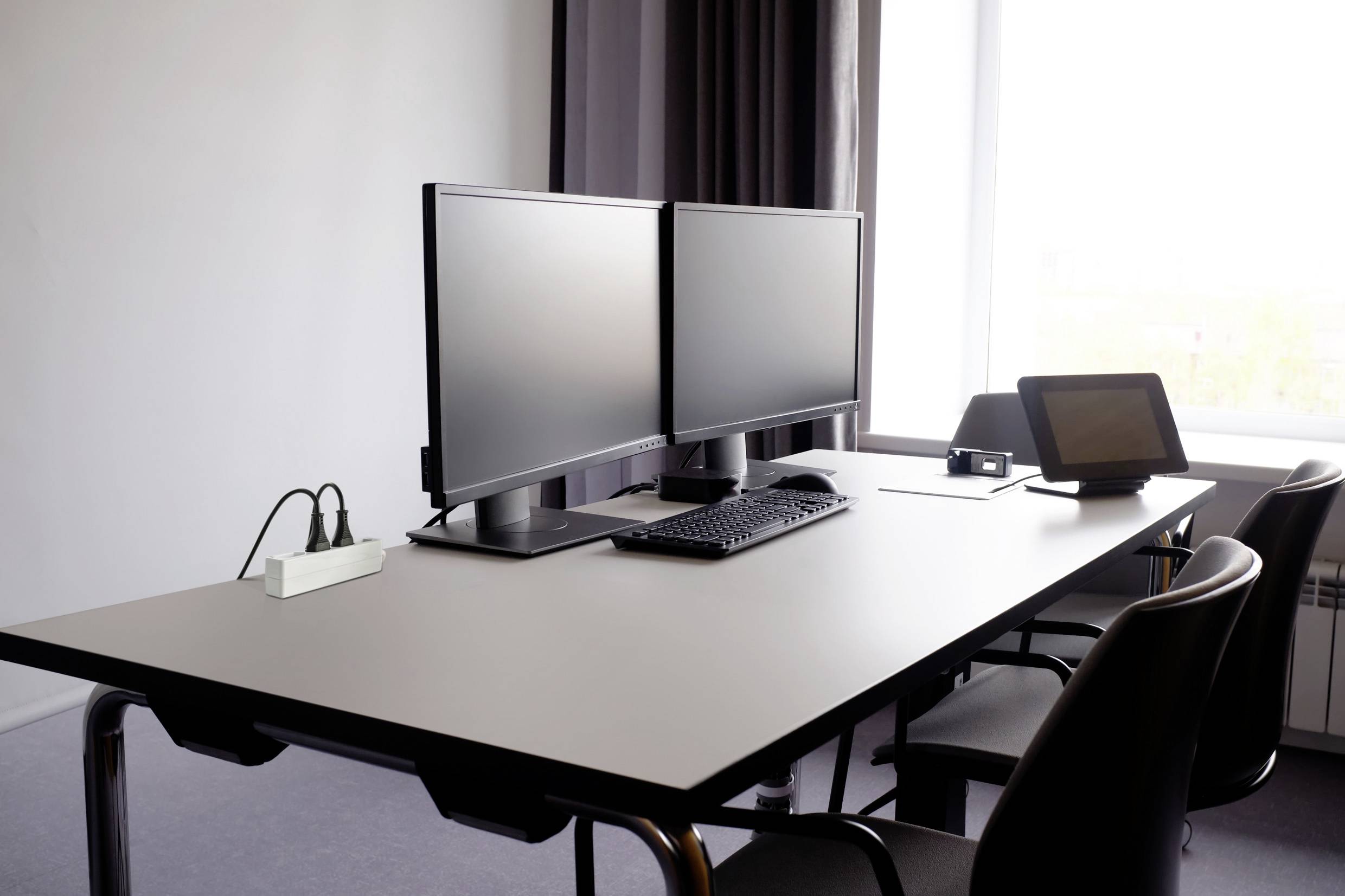 A conference room with a table, two monitors, a keyboard, a mouse and a tablet. A window can be seen in the background.
