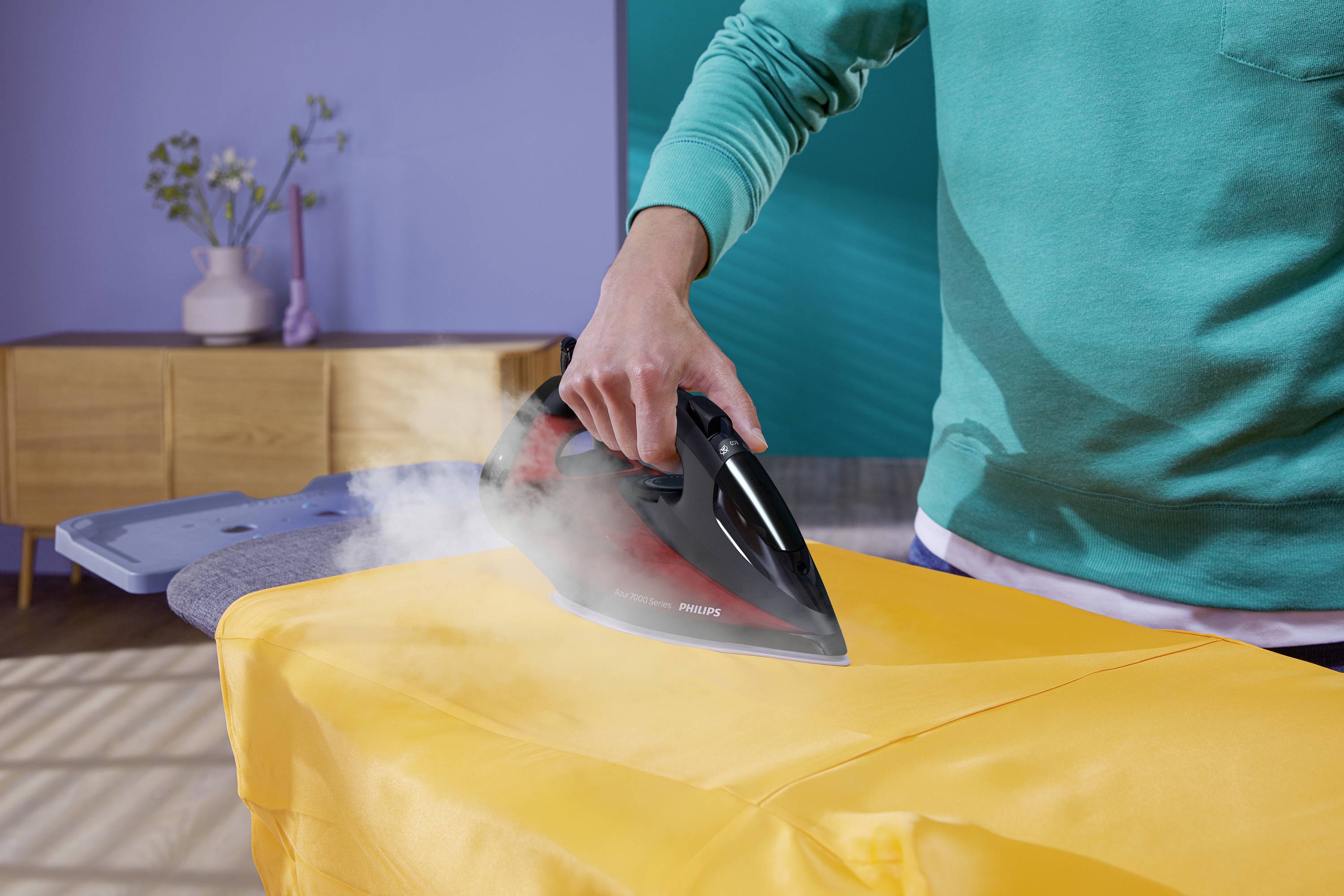 A person is ironing an orange shirt on an ironing board. In the background, there is a wooden piece of furniture with a flower vase.