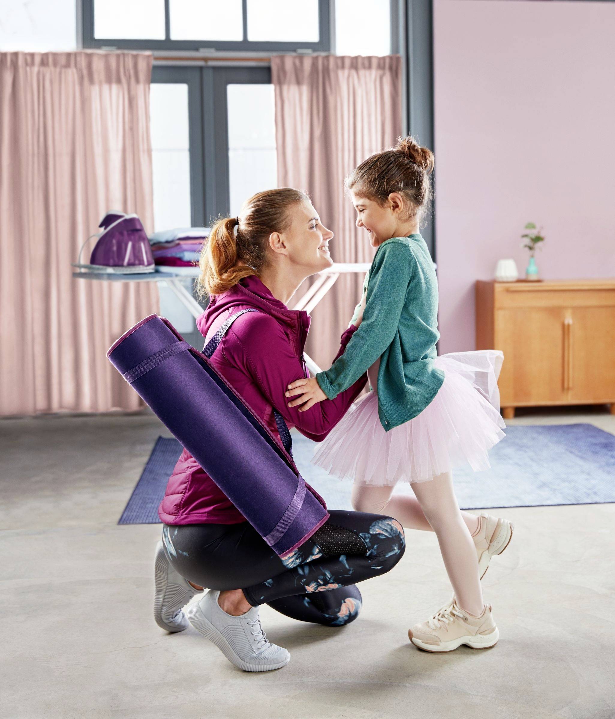 A smiling woman in sportswear kneels in front of a girl wearing a ballet costume and embraces her lovingly. Both appear happy. In the background, there is a window, an ironing board, and curtains.