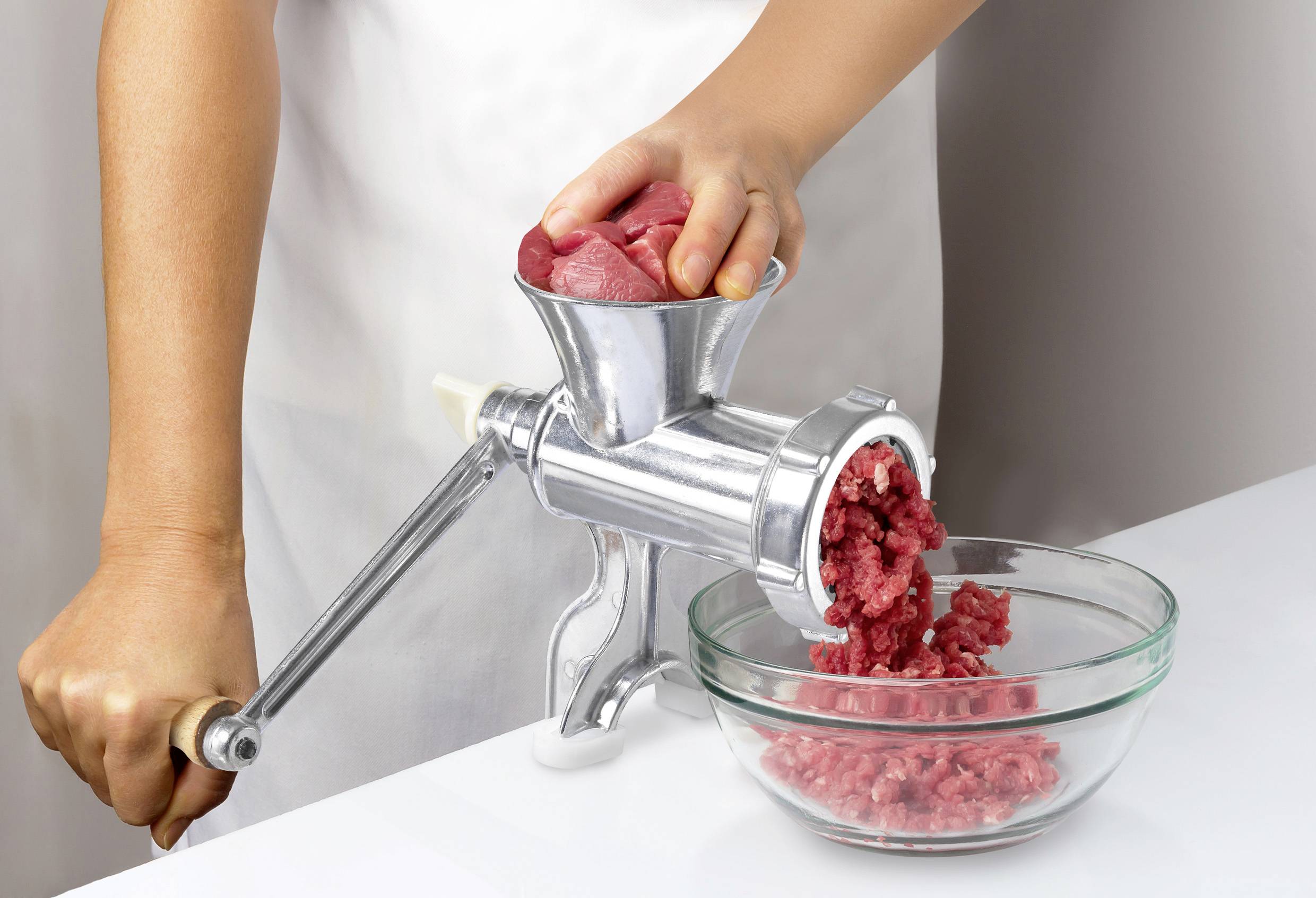 A person is mincing meat with a manual mincer, while the minced meat falls into a glass bowl.