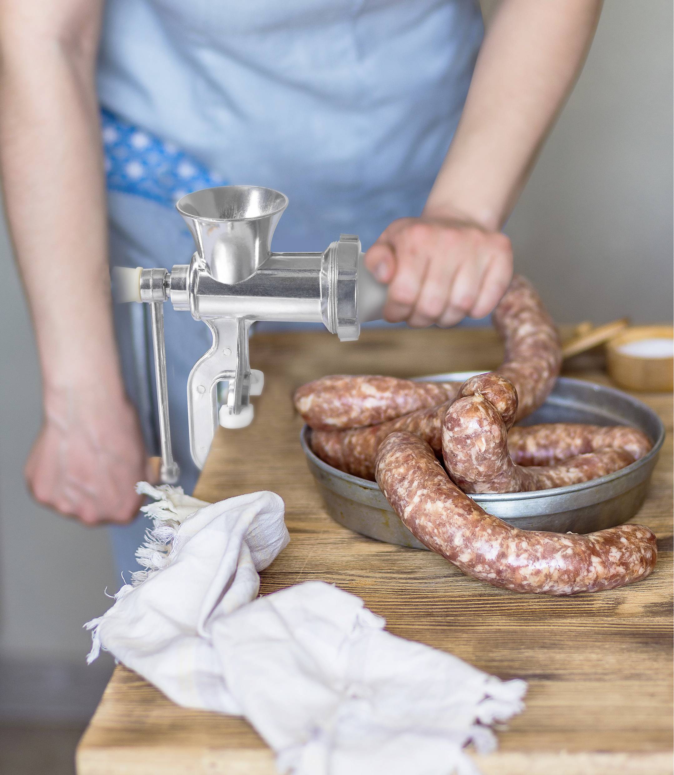 A person is filling sausage mixture into a manual mincing machine on a wooden table. Two raw sausages are lying in a metal container.
