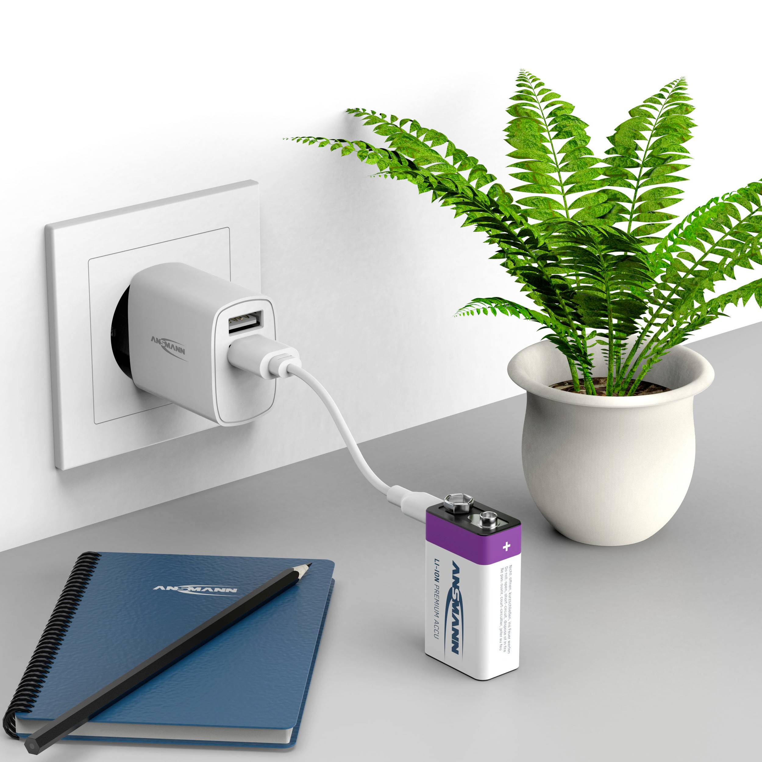 Potted plant next to a charger charging a battery. Charger plugged into a socket. Notebook lying on the table.