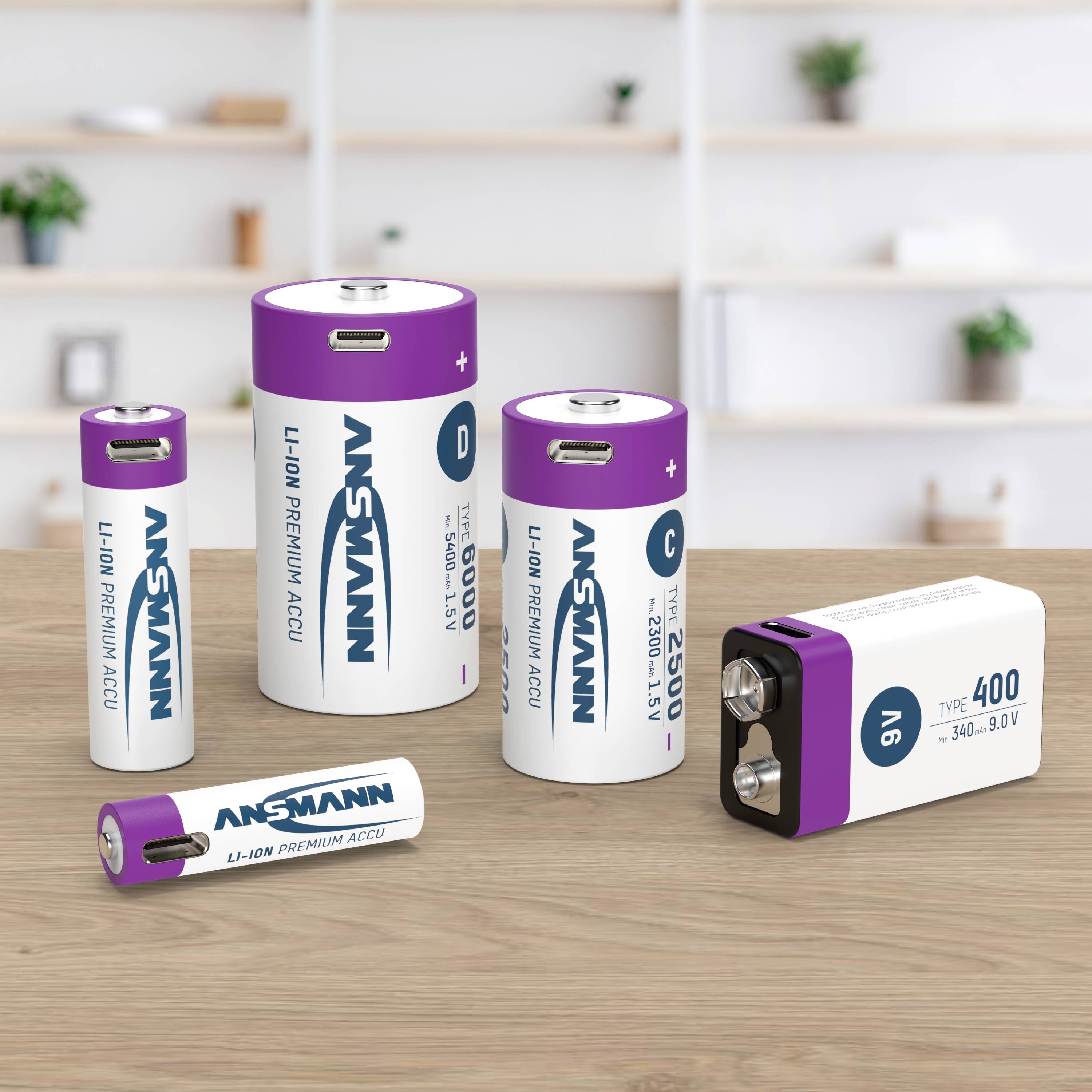 Five different Ansmann batteries on a table. In the background, blurry shelves with plants and objects.