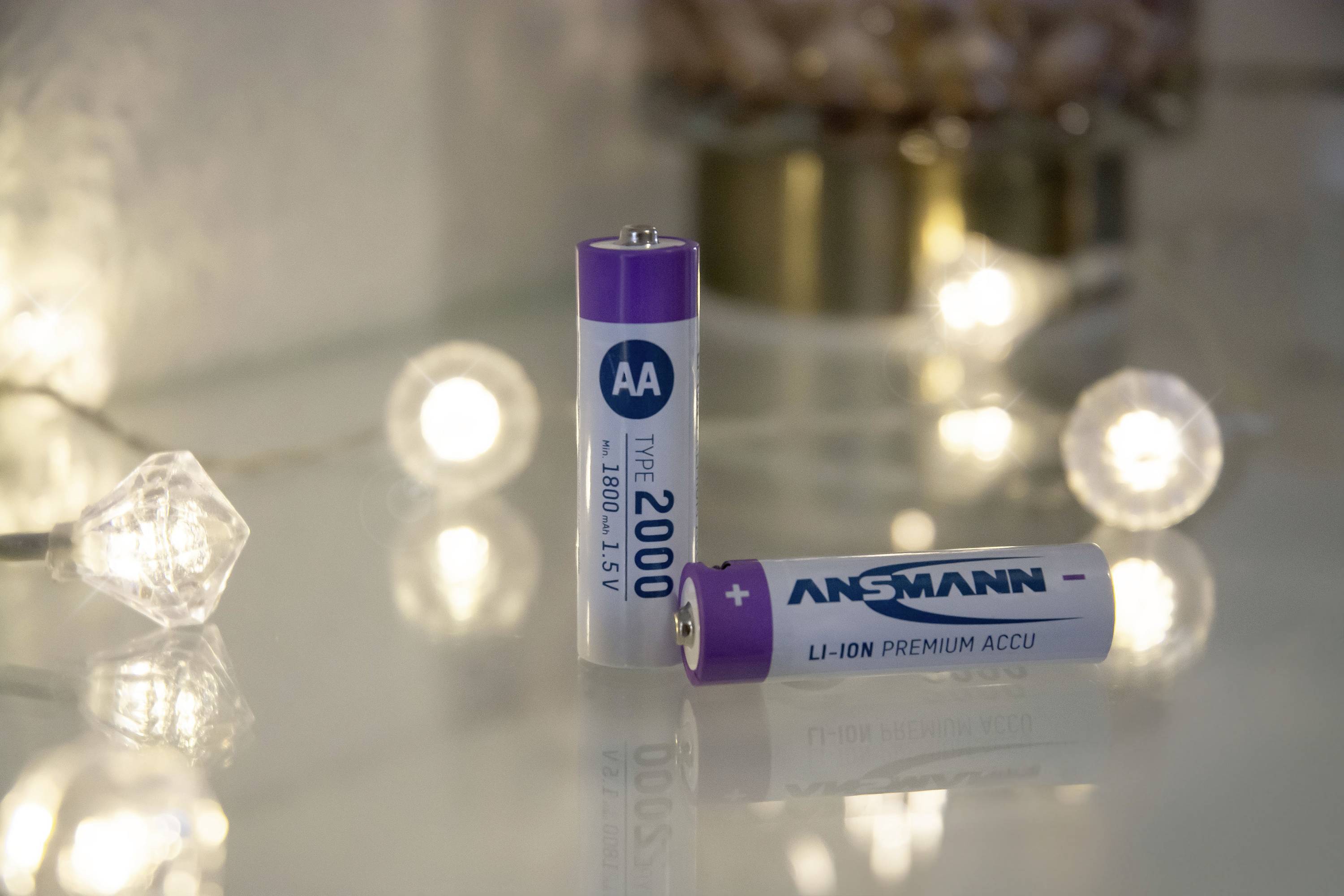 Two AA batteries, one standing and one lying down, on a table. In the background, blurred fairy lights are visible.