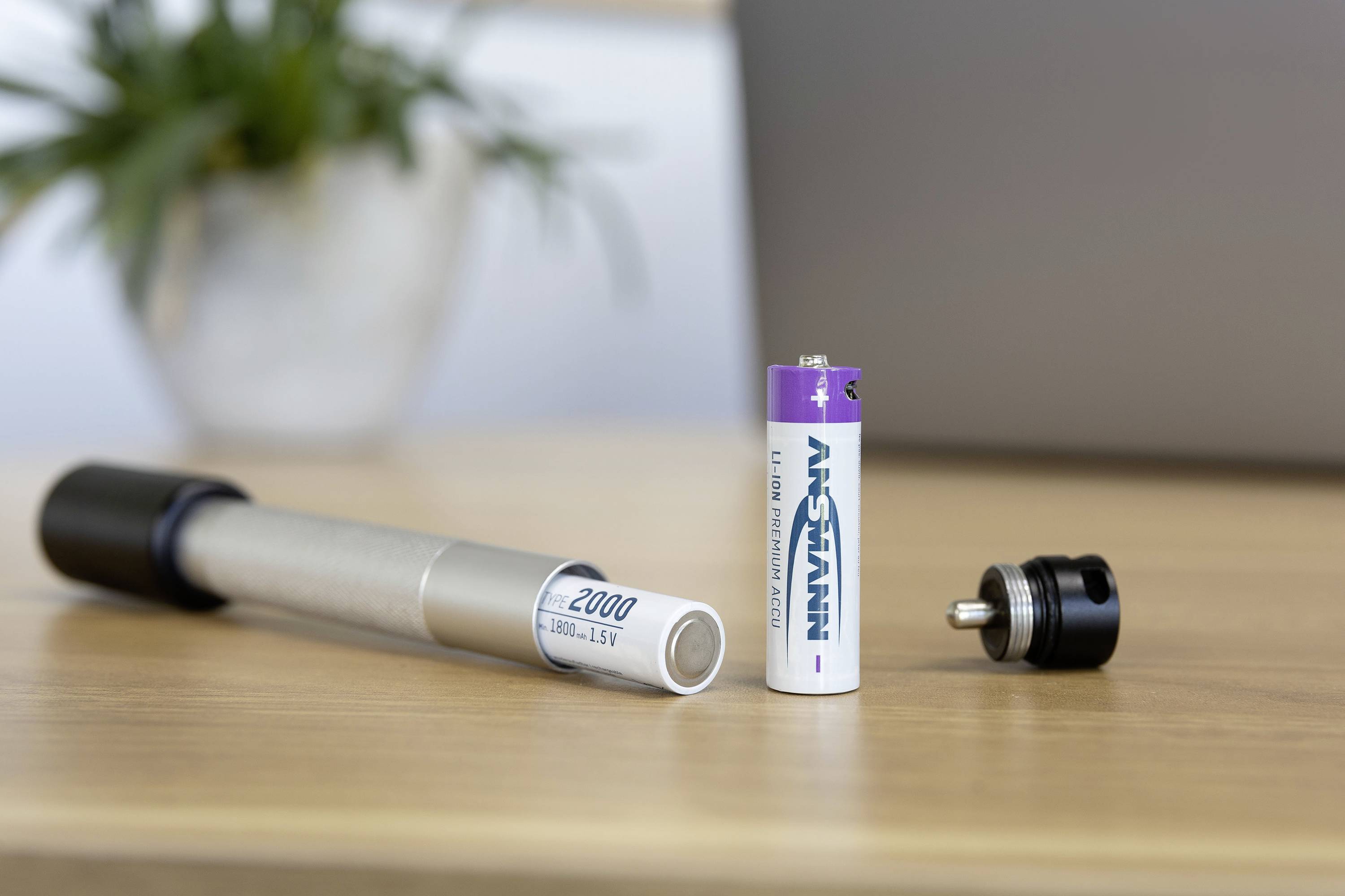 A battery and a part of a torch rest on a wooden table. A plant is slightly out of focus in the background.