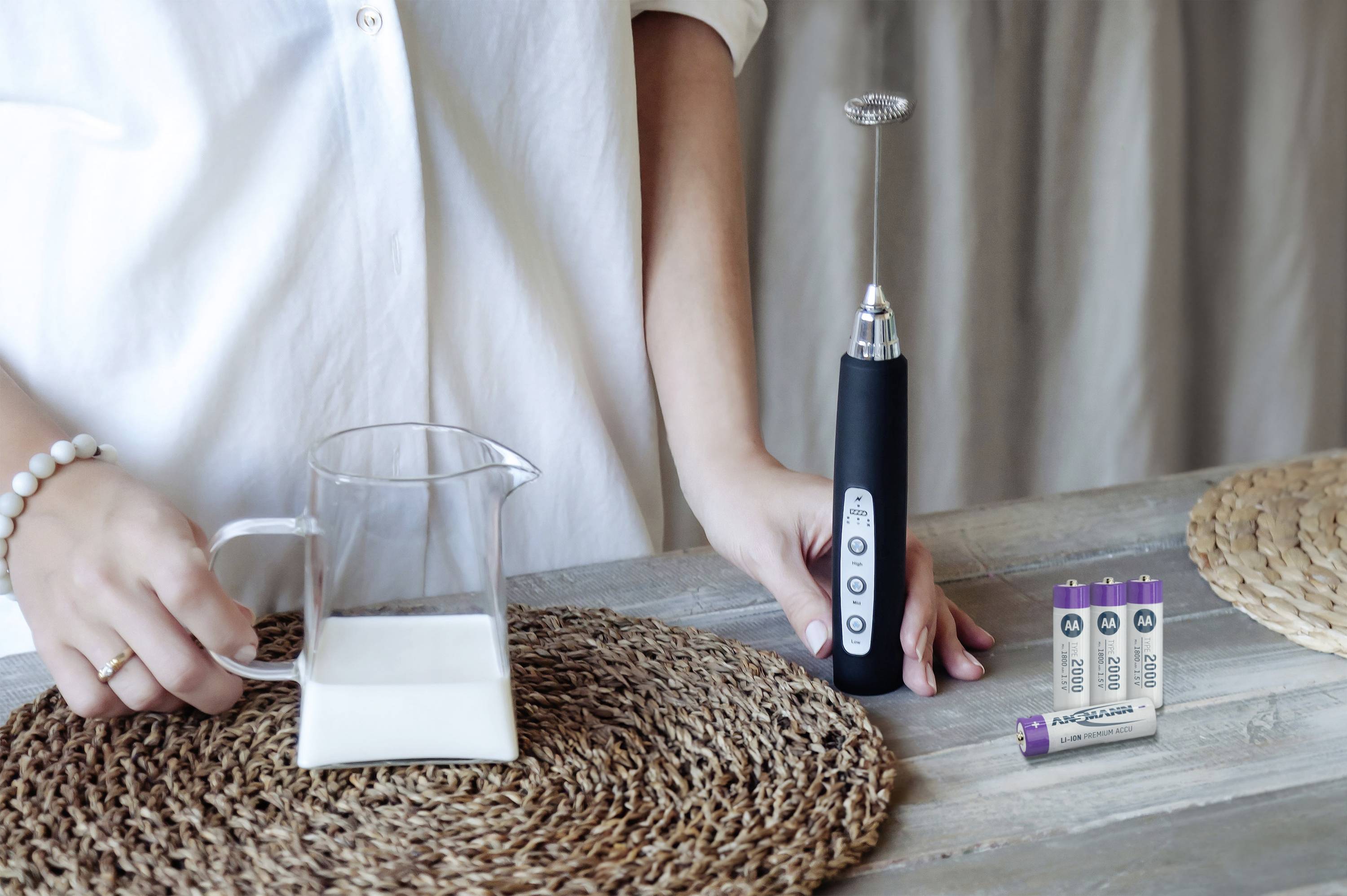 A person is holding a milk frother next to a milk jug and batteries on a table.