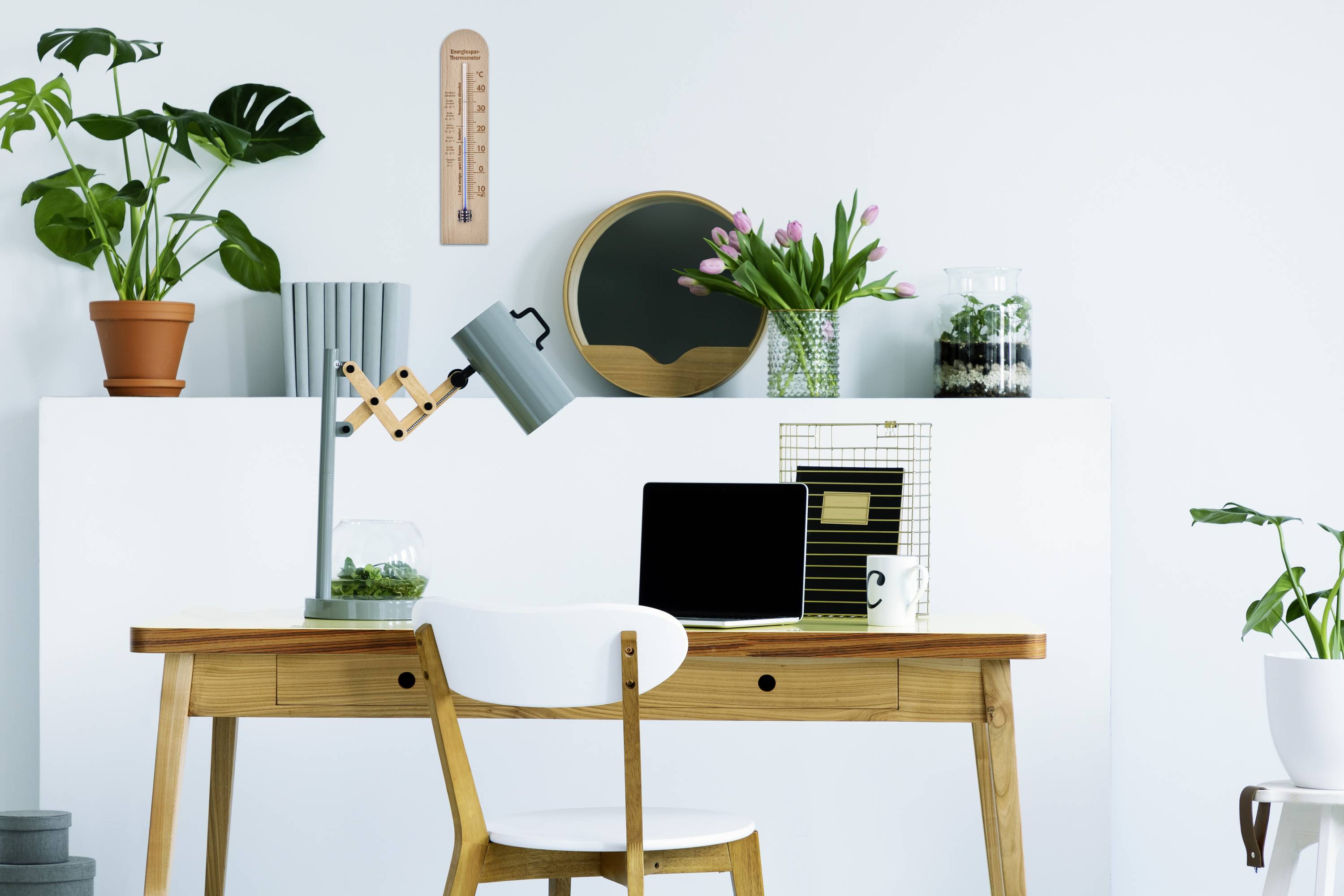 Home office with a wooden desk, laptop, watering can as a lamp, plants in the background. Minimally and modernly furnished.