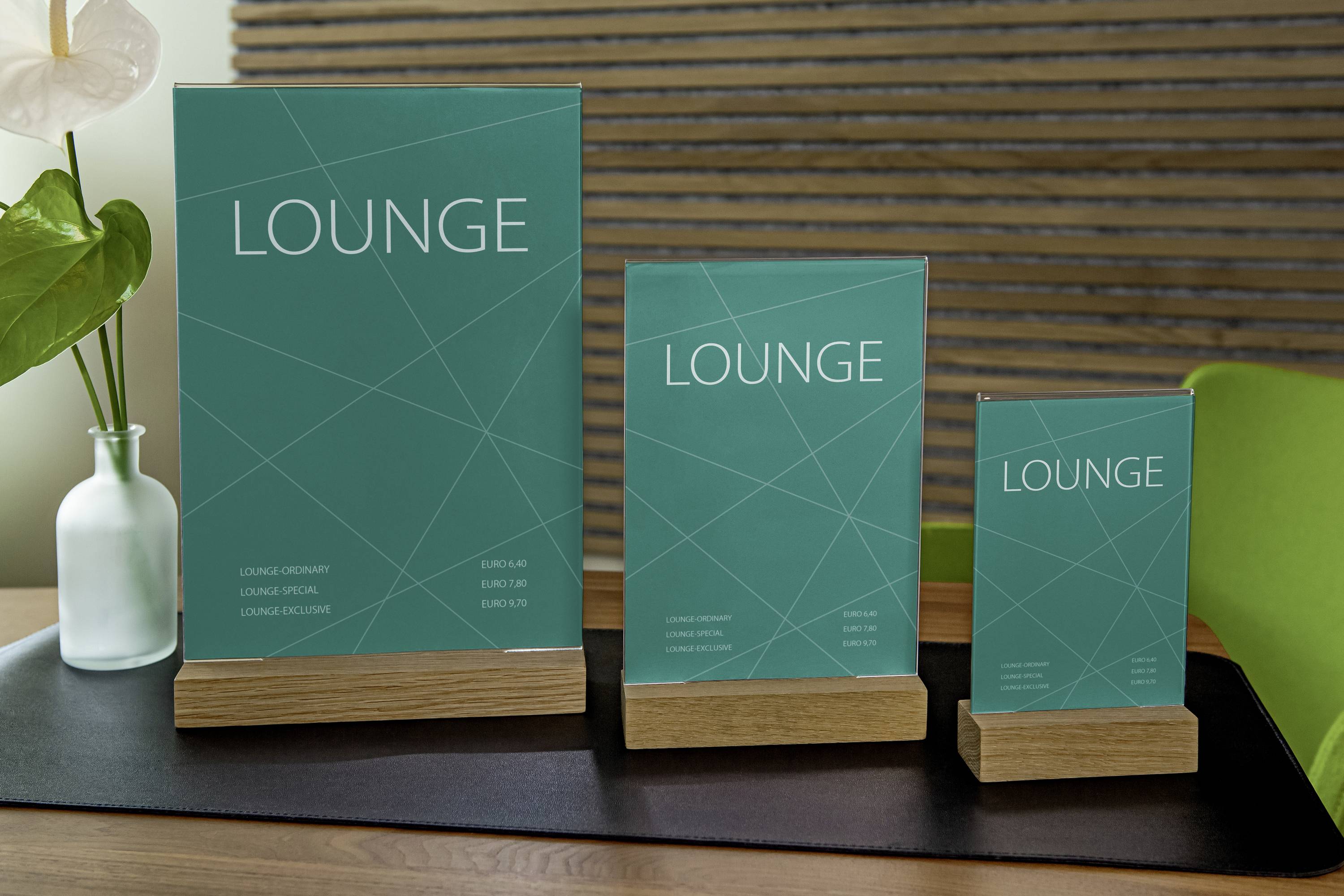 Three green signs in wooden stands with the inscription 'LOUNGE'. Different sizes, placed on a table with plants in the background.