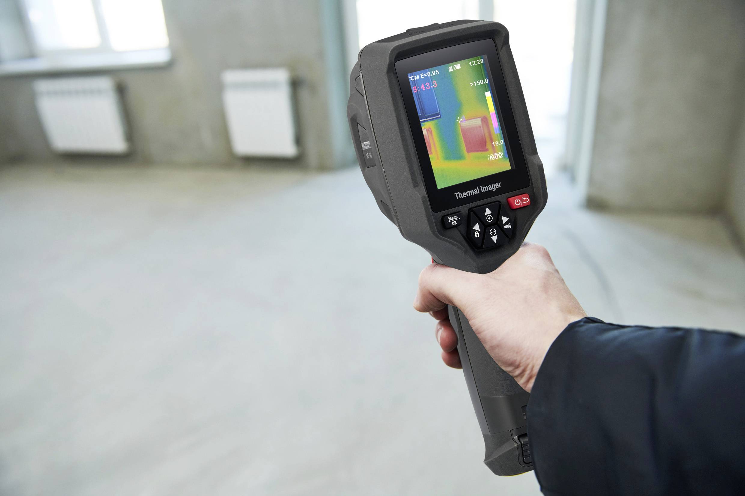A person is holding a thermal imaging camera in an empty room, displaying temperature variations on a screen.