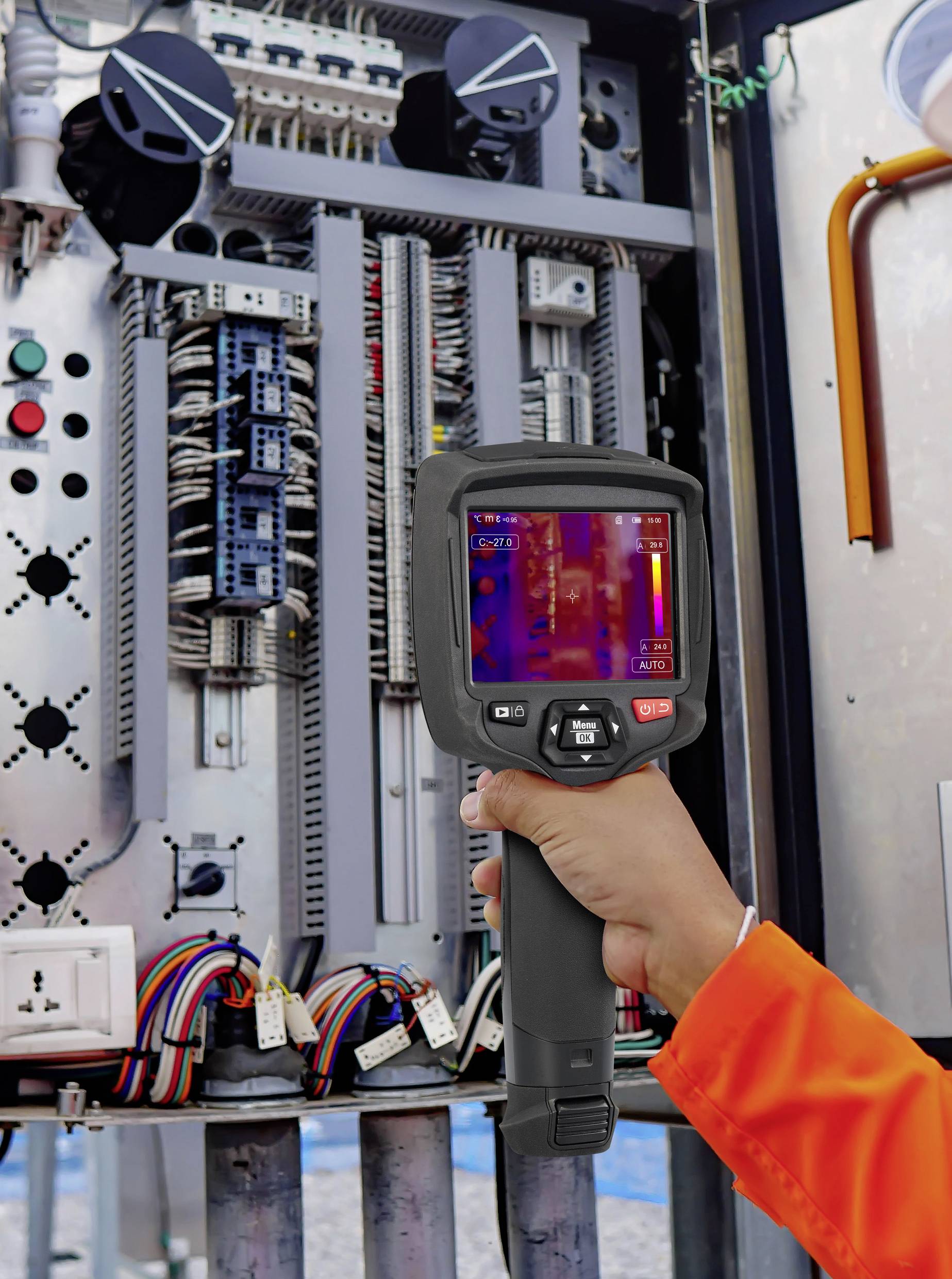 A person is holding a thermal imaging camera in front of an electrical control cabinet. The screen displays temperature variations.