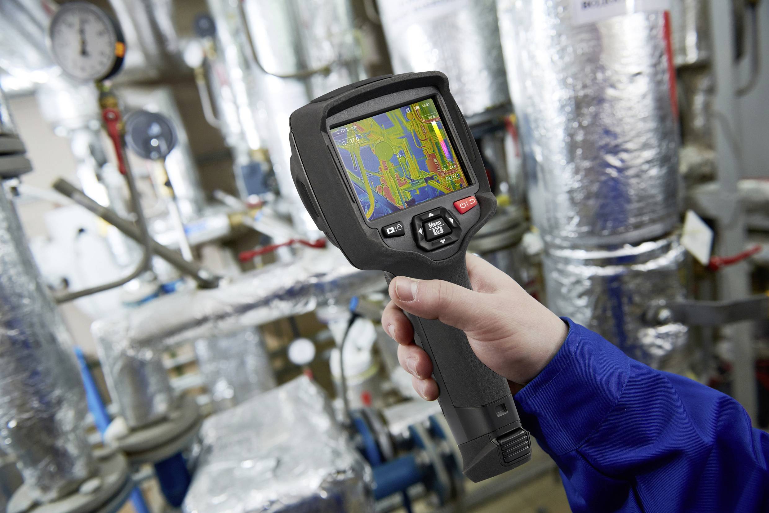 A person is holding a thermal imaging camera, checking pipework in a machinery room. Coloured temperature zones are visible on the display.
