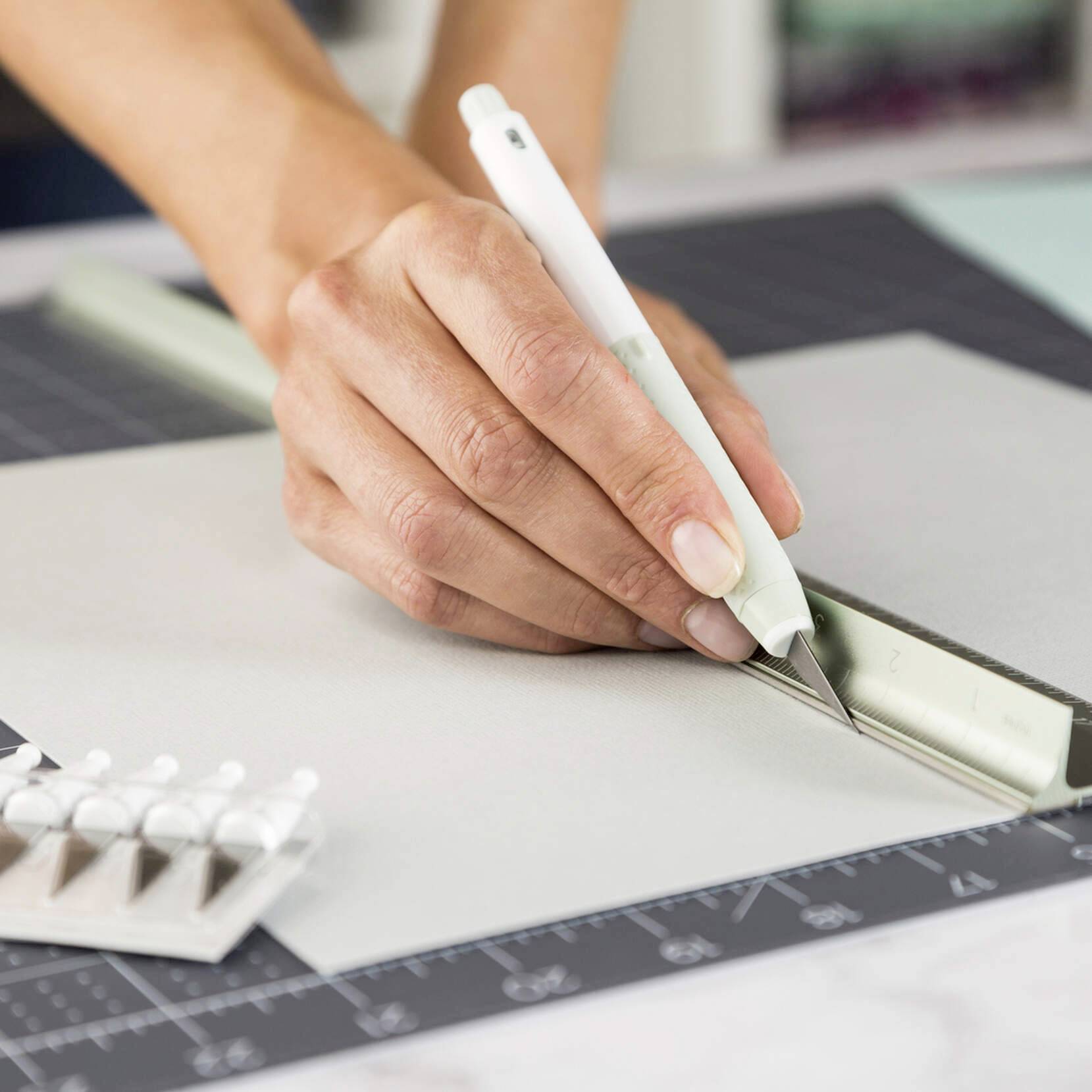 A hand is holding a white cutting tool, slicing paper along a metal edge on a cutting mat.