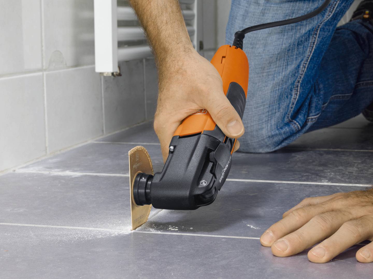 A person is repairing a tiled floor using an orange multi-functional electric tool. The tool is cutting through the grout.