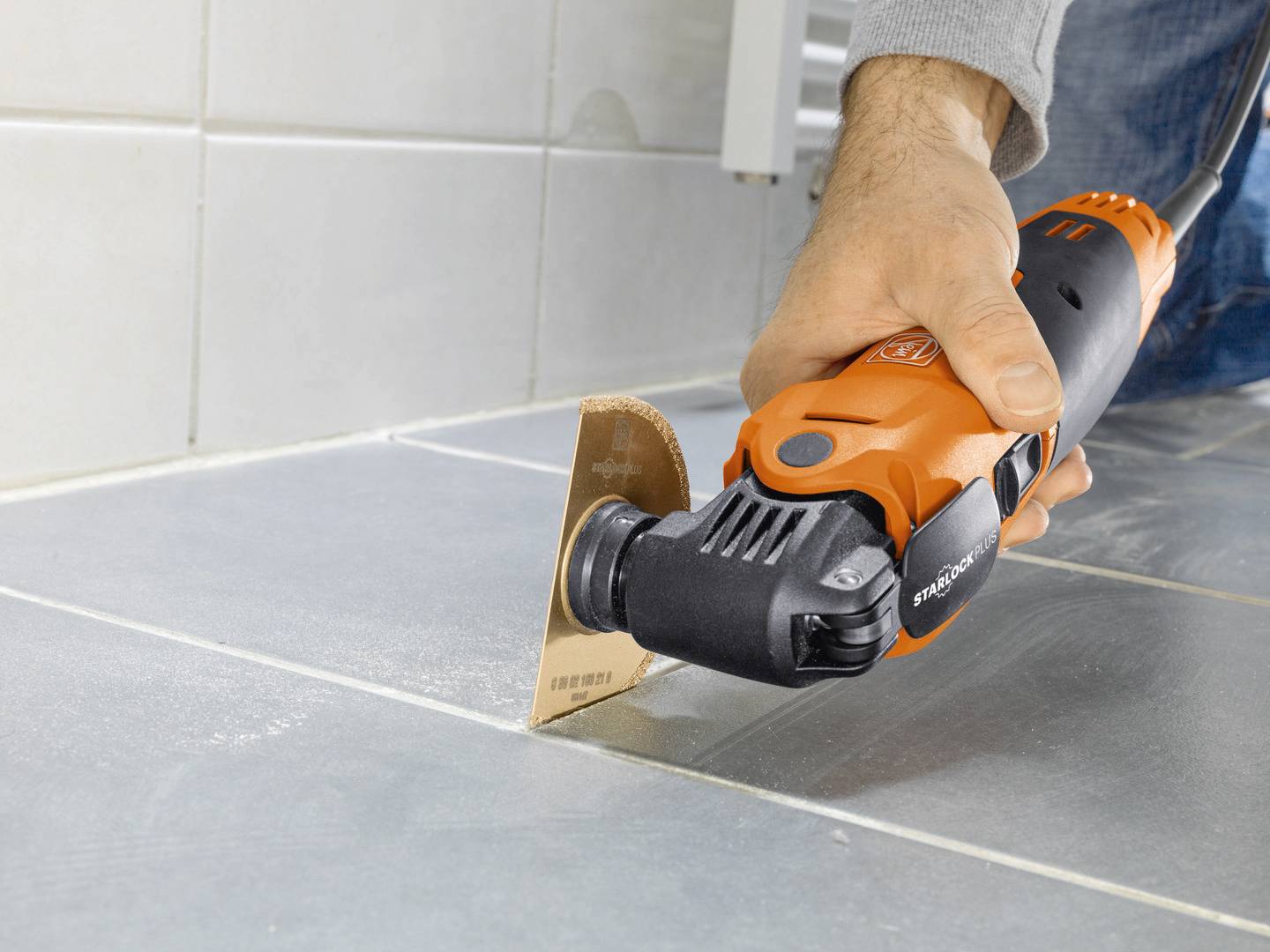 A person is using an oscillating tool to remove grout between grey tiles.