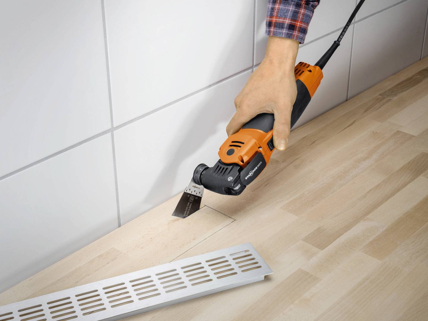 A person is precisely cutting a rectangular opening in a wooden surface using an oscillating multi-tool. A ventilation grille is lying beside it.