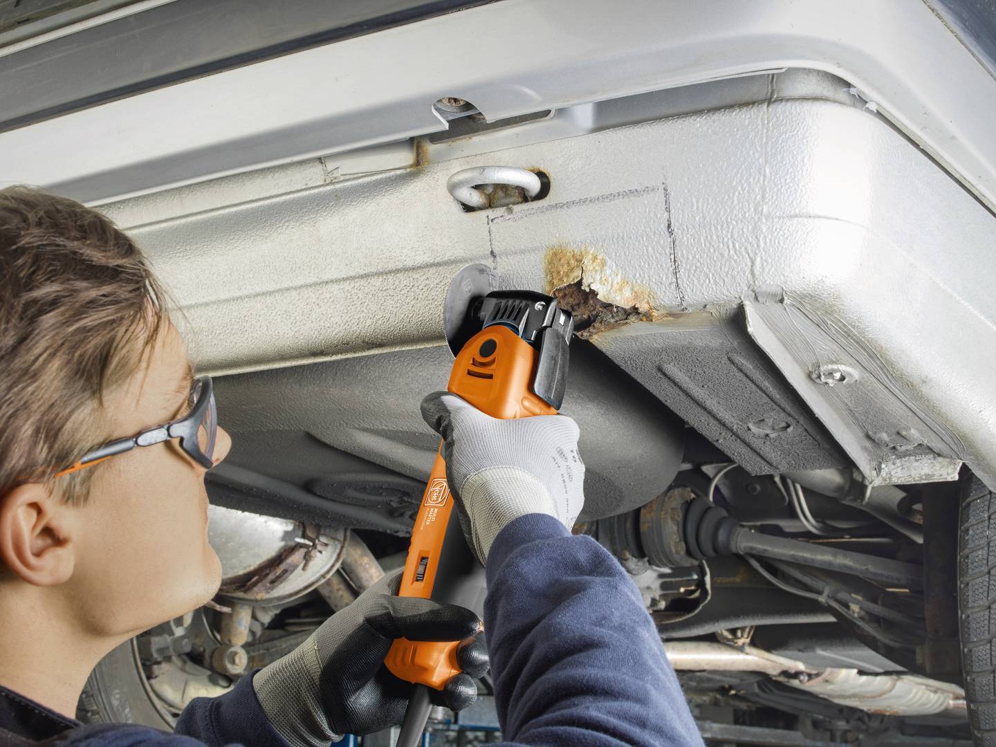 A person is removing rust from the underside of a vehicle using an electric tool. Safety glasses and gloves are being worn.