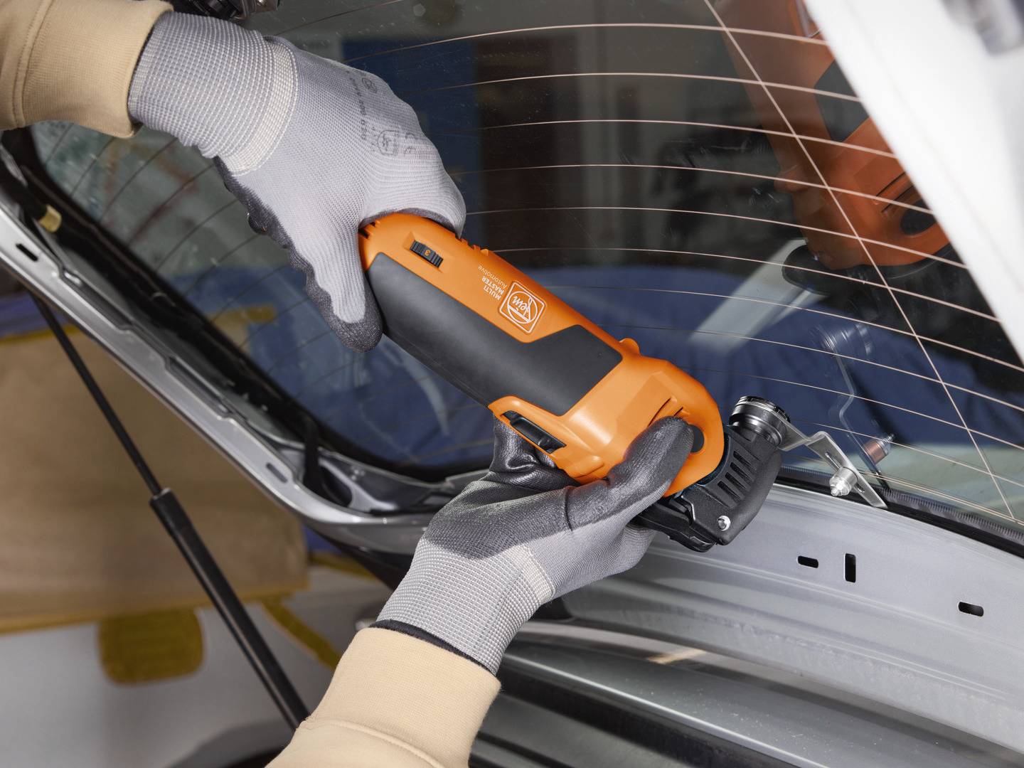 A person is using an orange electric tool to work on the rear windscreen of a car. They are wearing grey gloves.