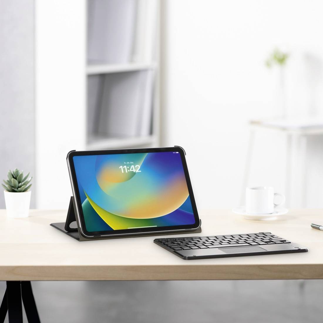 A tablet in a case sits on a table next to a keyboard. Blurred shelves and a mug are visible in the background.