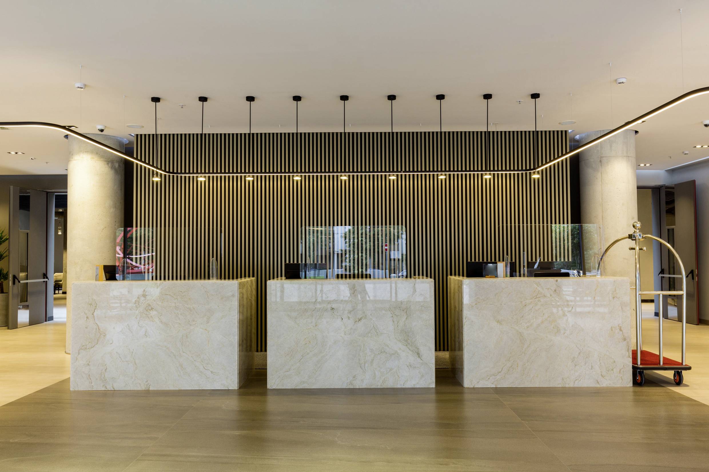 Reception of a hotel with three marble counters, modern ceiling lights, and an elegant design featuring wooden slats in the background.