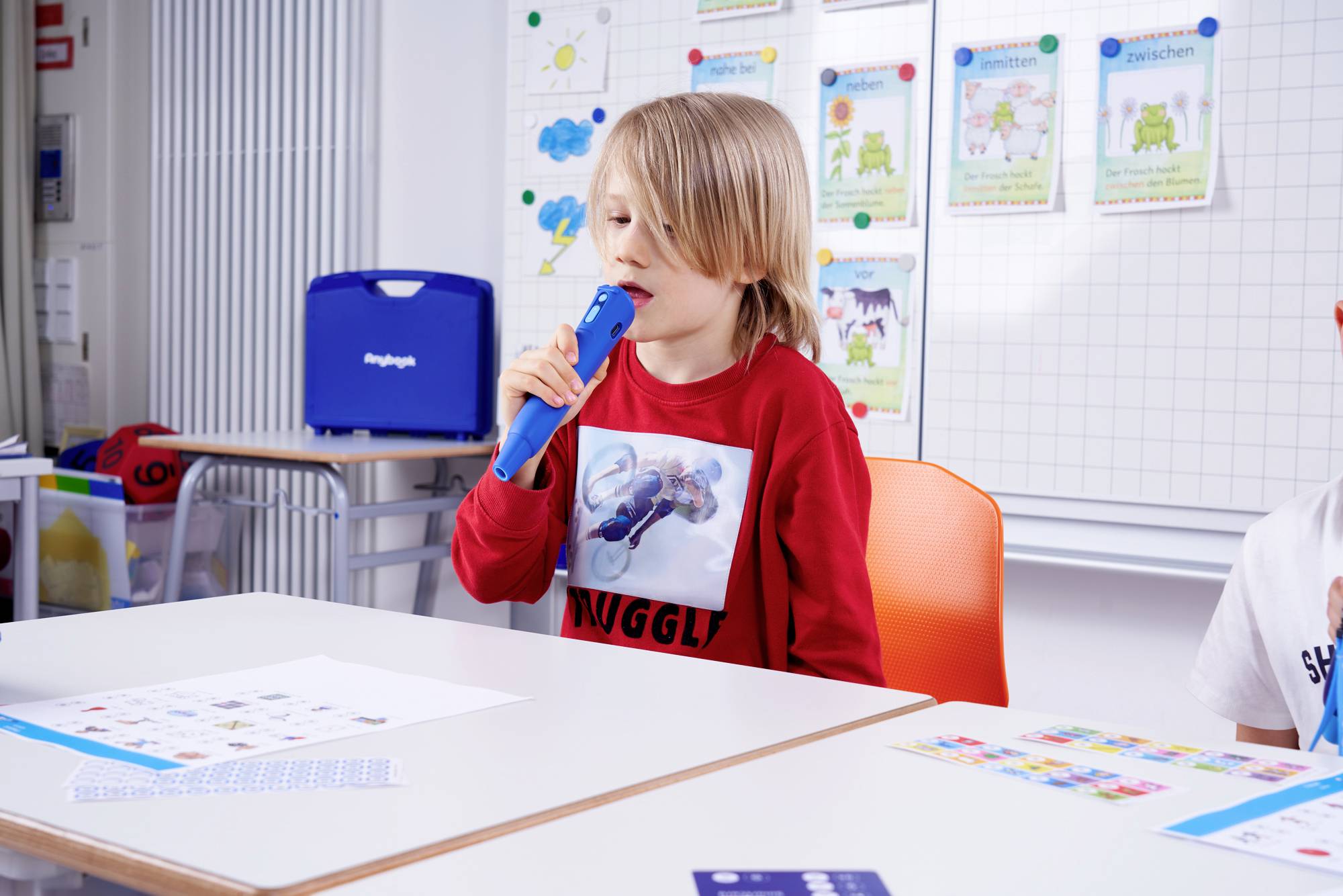 A child in a classroom is using an electronic reading device. In the background are colourful learning posters and materials.