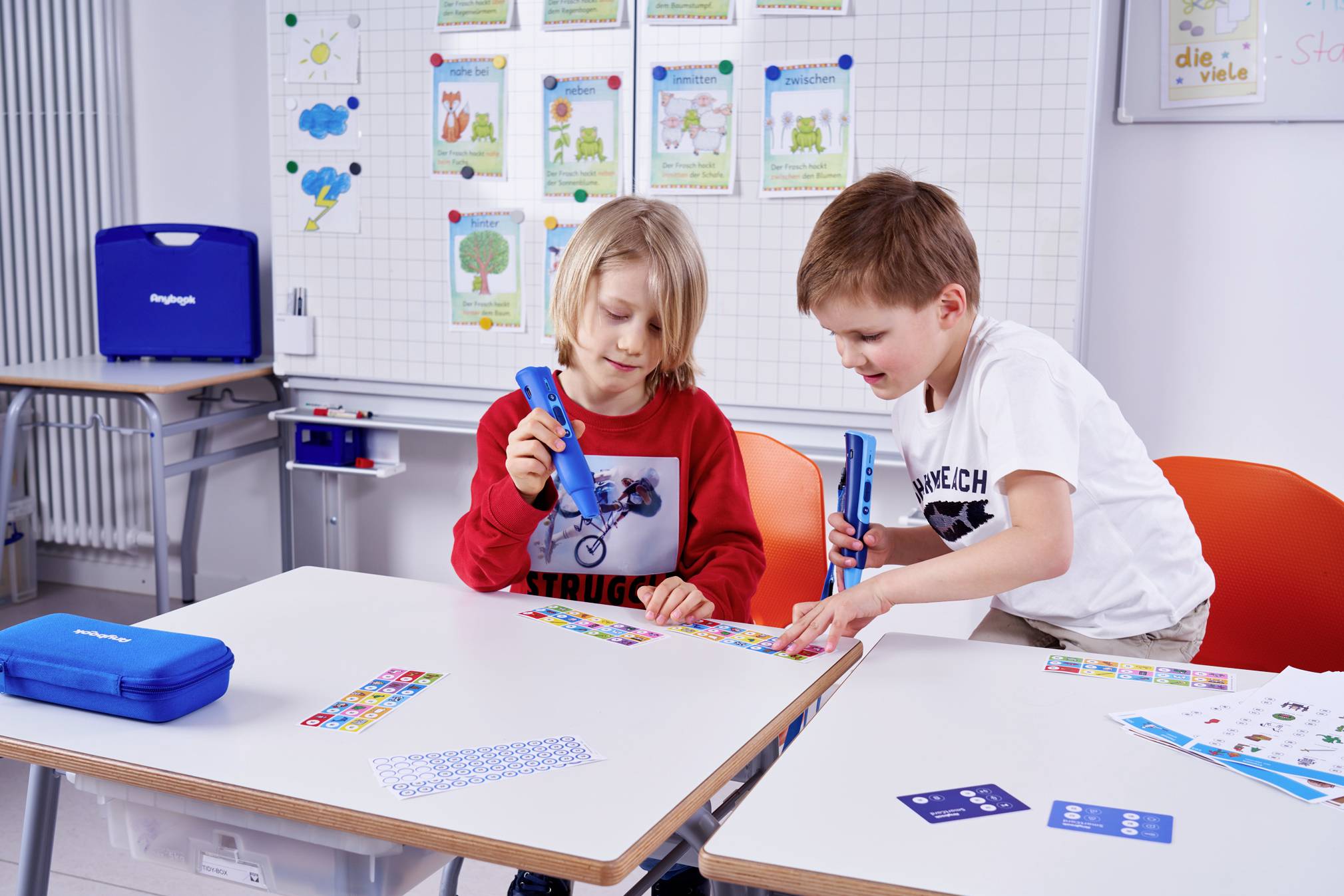 Two children are sitting at a table in a classroom, working with colourful stickers. Learning posters are displayed on the wall in the background.