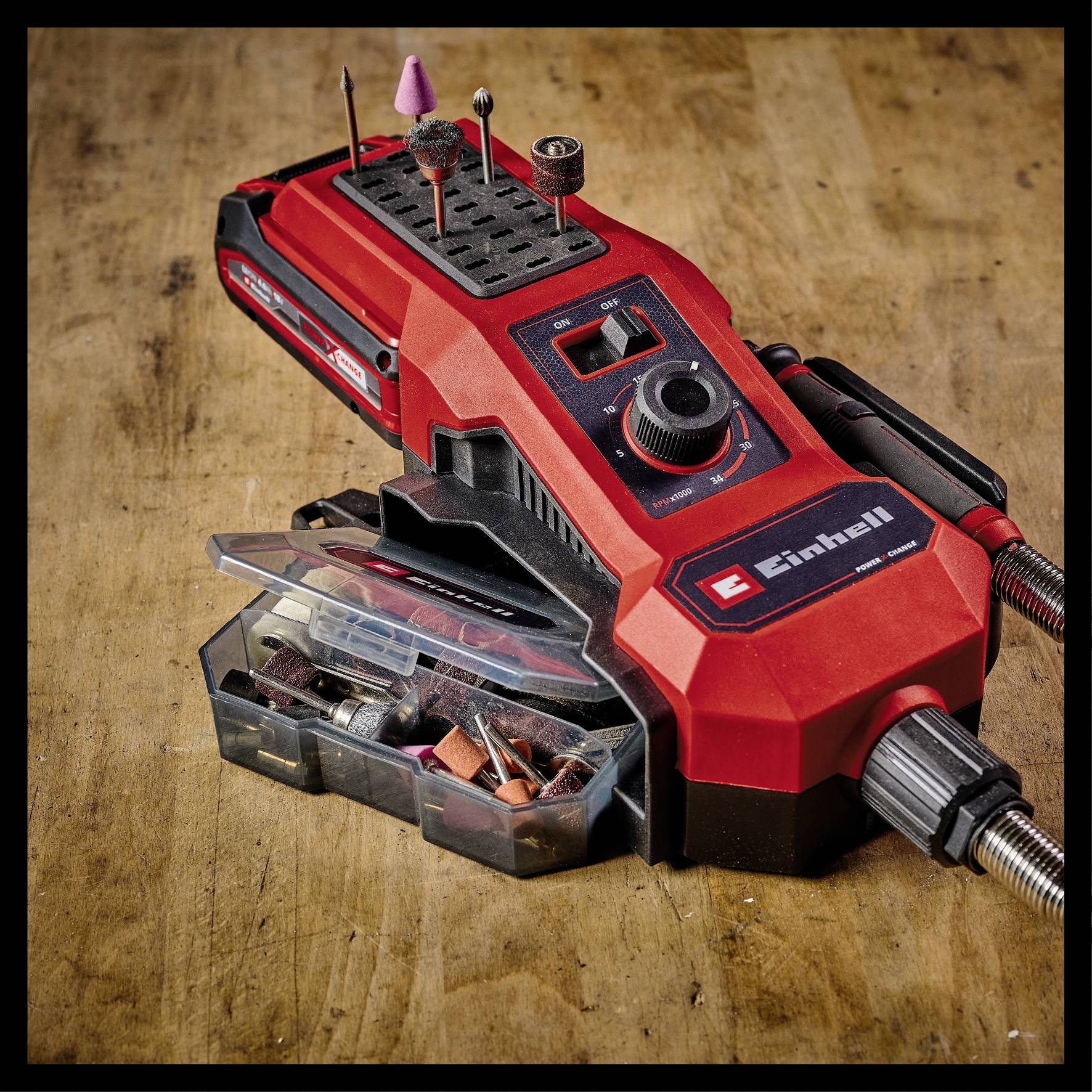 A red, portable multi-tool with various drill and grinding attachments on a wooden table. Accessories are stored in a clear box.