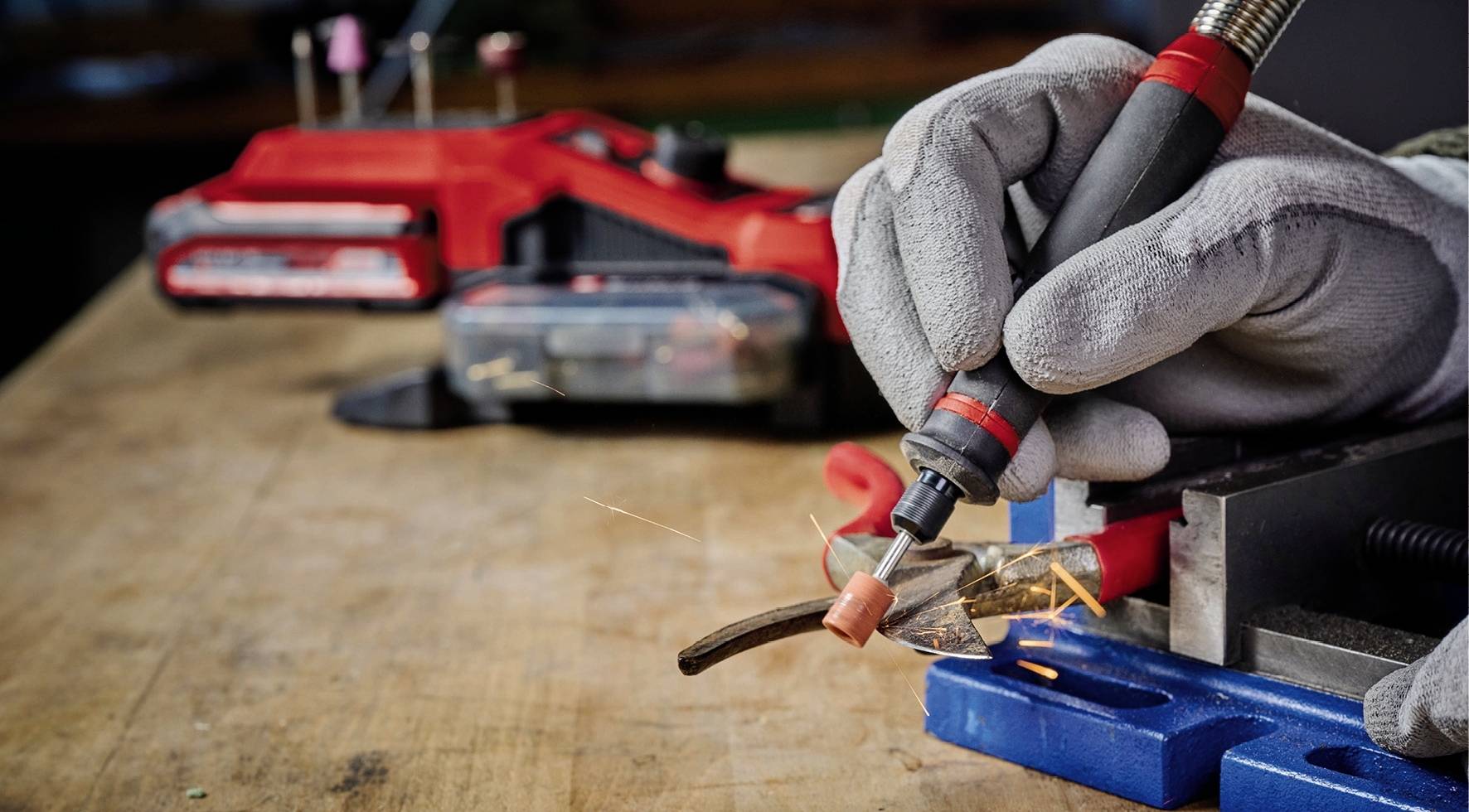 A person wearing gloves is carrying out precise soldering on a metal piece on a workbench. A red tool is visible in the background.
