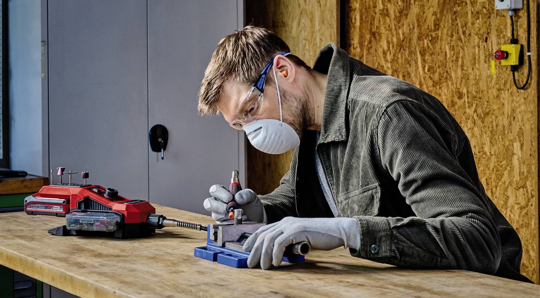 A man is working intently at a table, wearing a protective mask and gloves, and using a tool for precise manual work.