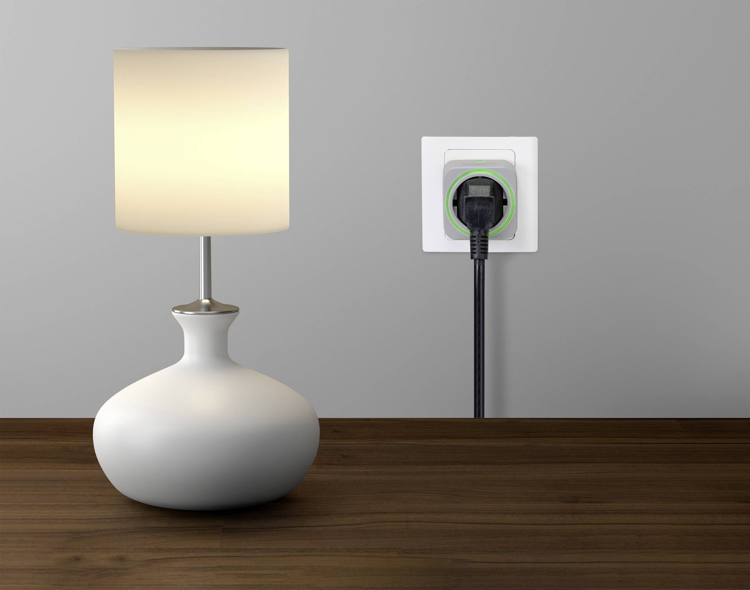 A switched-on table lamp sits on a wooden table; beside it a power socket with a plugged-in cable.