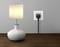 A switched-on table lamp sits on a wooden table; beside it a power socket with a plugged-in cable.