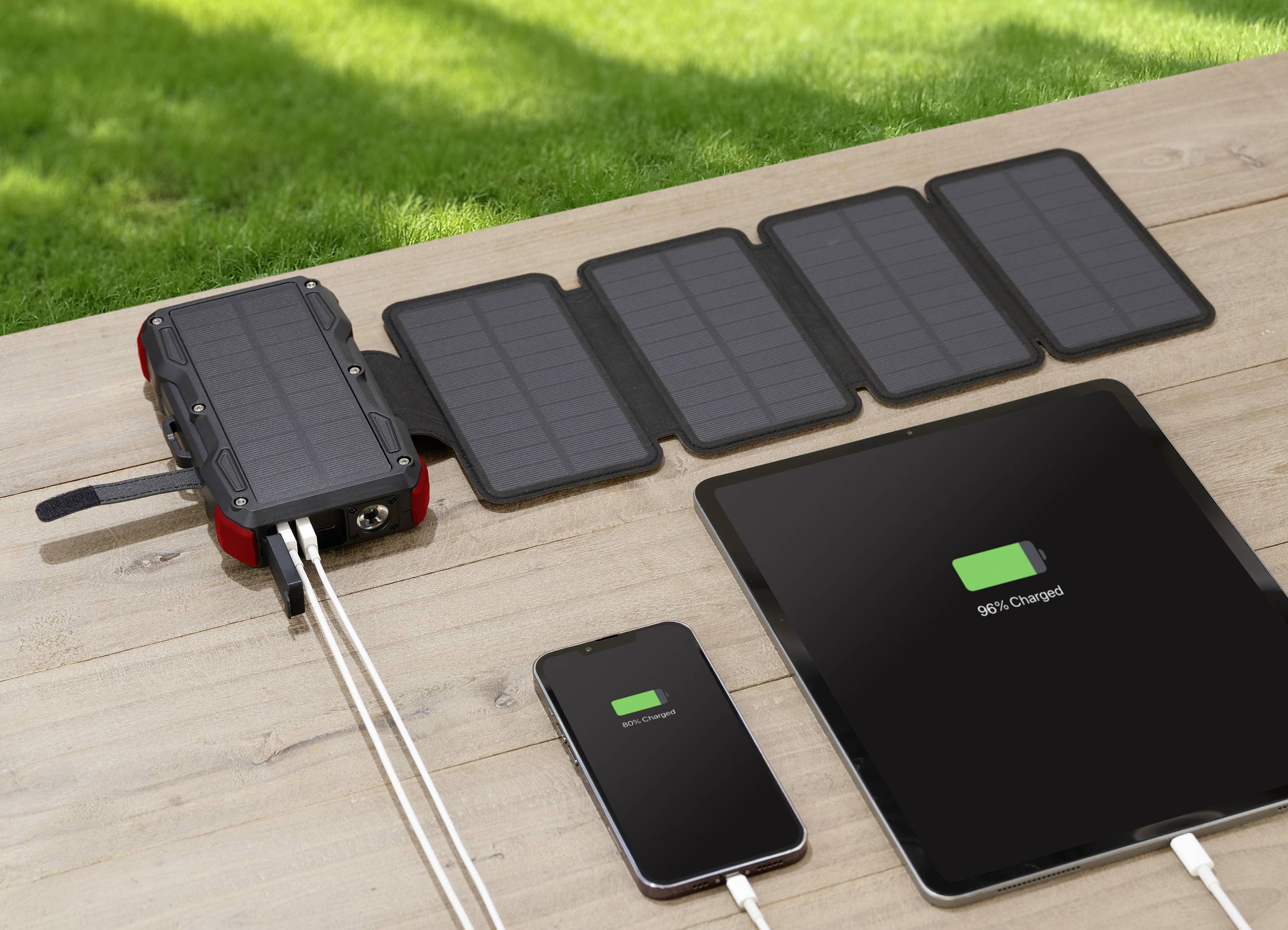 Solar-powered chargers on a wooden table are charging a smartphone and a tablet. Both devices are showing 98% and 99% 'charged'.
