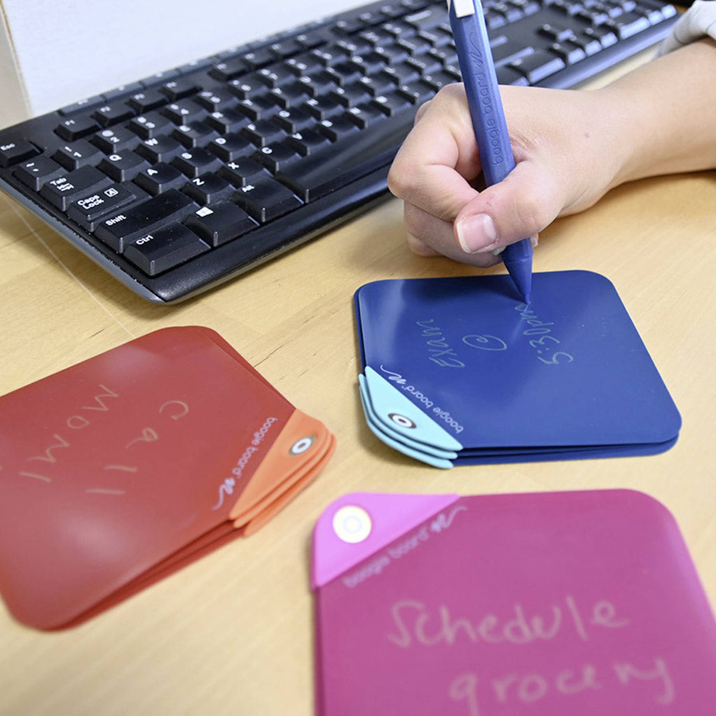 A hand is writing with a pen on a blue sticky note. Coloured notes are lying on the table. A computer keyboard is visible in the background.