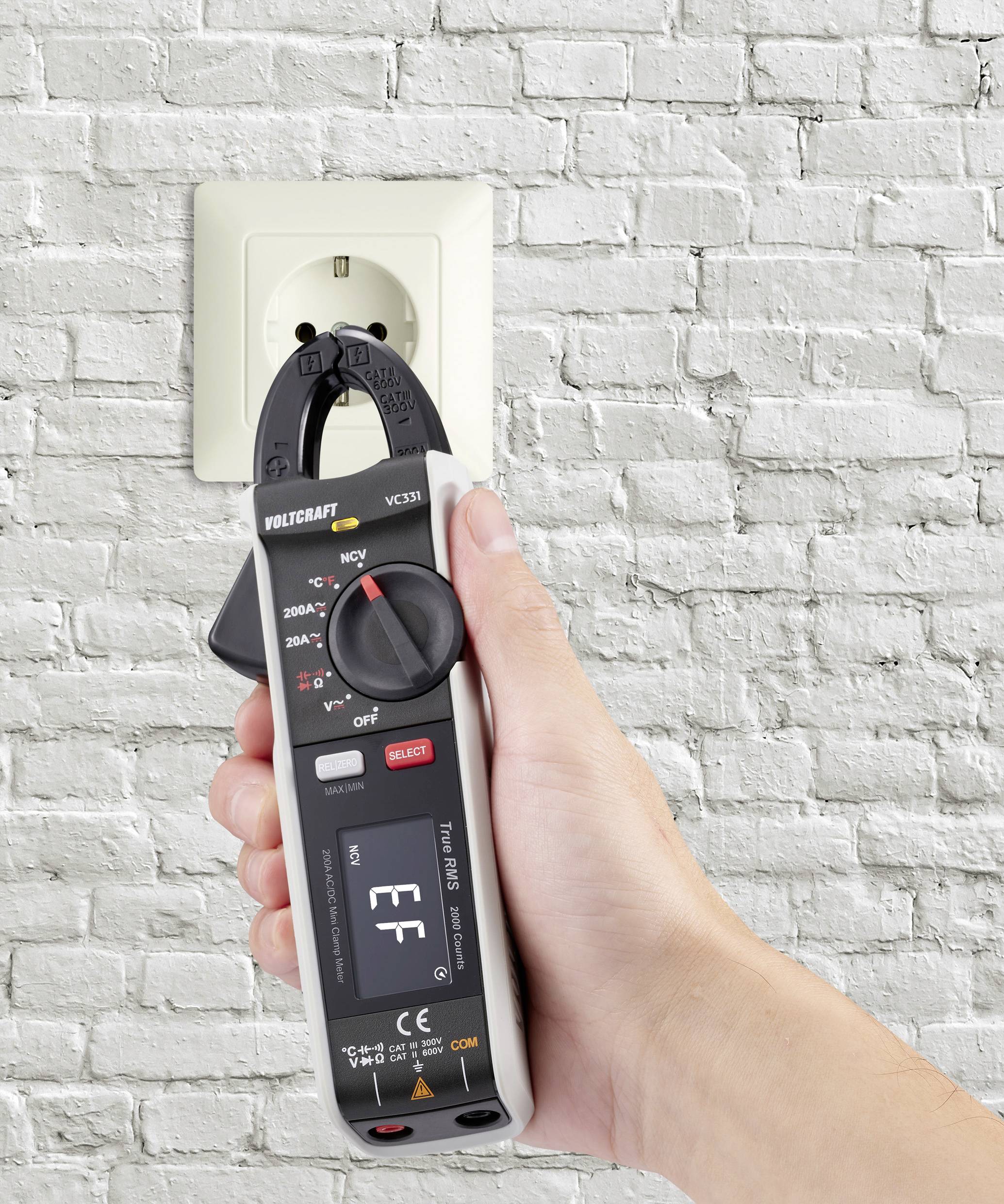 A hand holds a voltage tester in front of a white socket in a brick wall to measure the voltage.