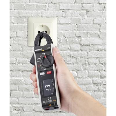 A hand holds a voltage tester in front of a white socket in a brick wall to measure the voltage.