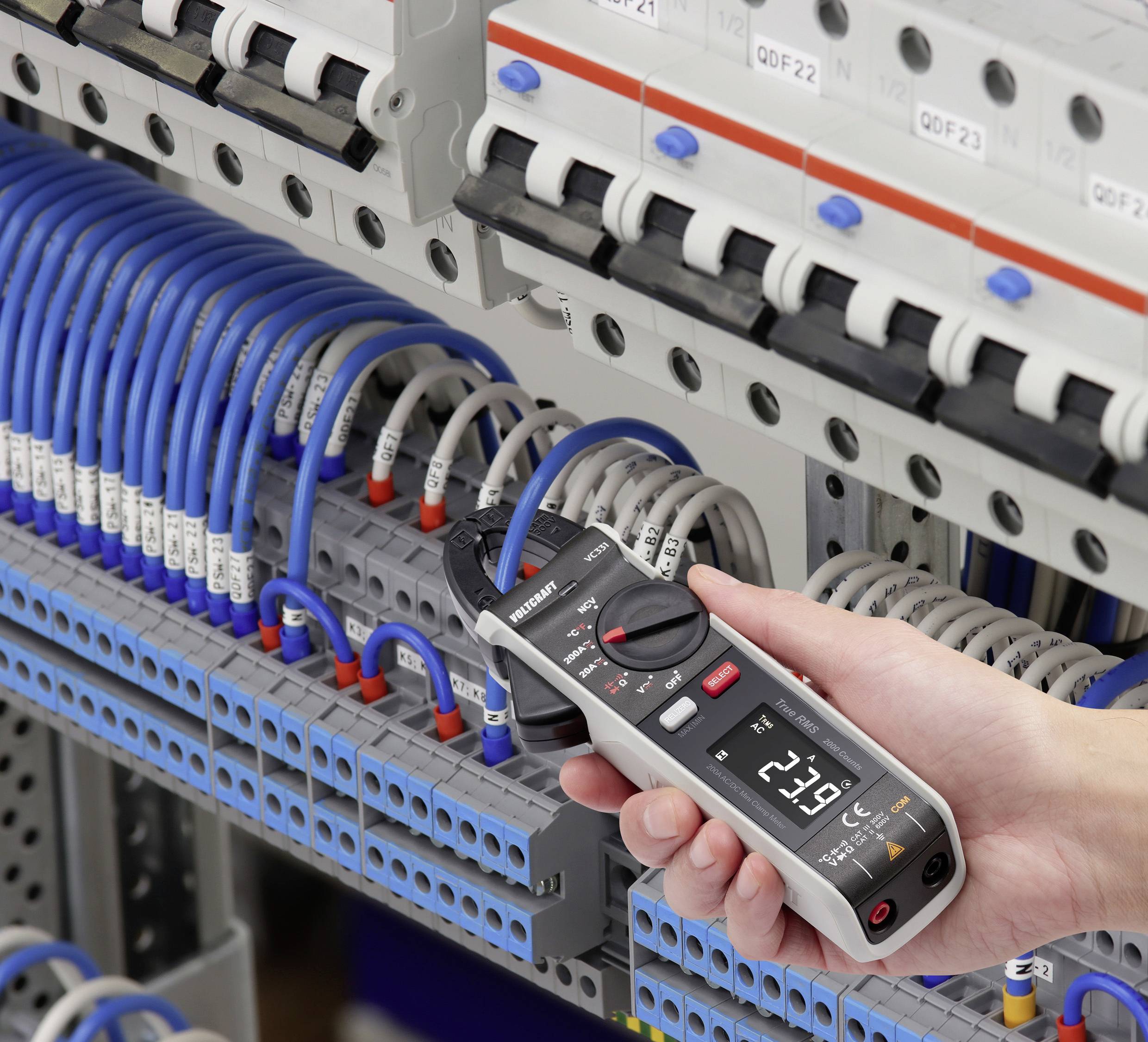 A person is measuring the voltage in an electrical cabinet with a digital multimeter. Cables are connected to various terminals.