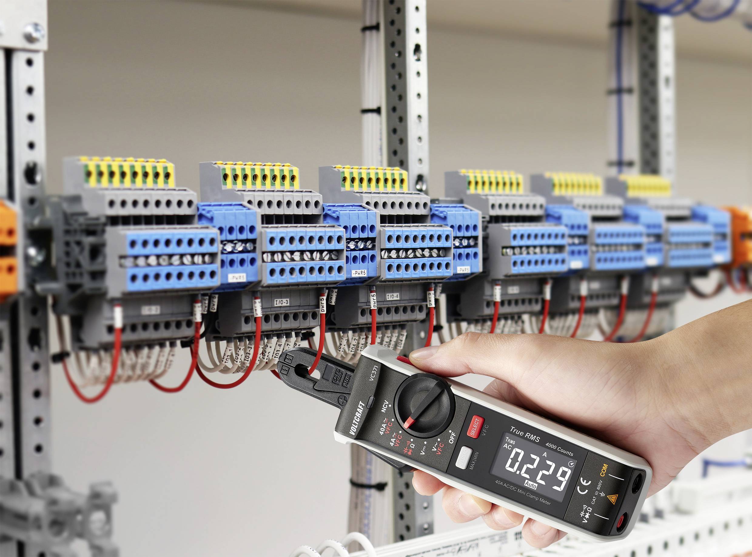 A hand is holding a digital measuring device connected to an electrical cabinet. The display shows '0.2239' while taking measurements.