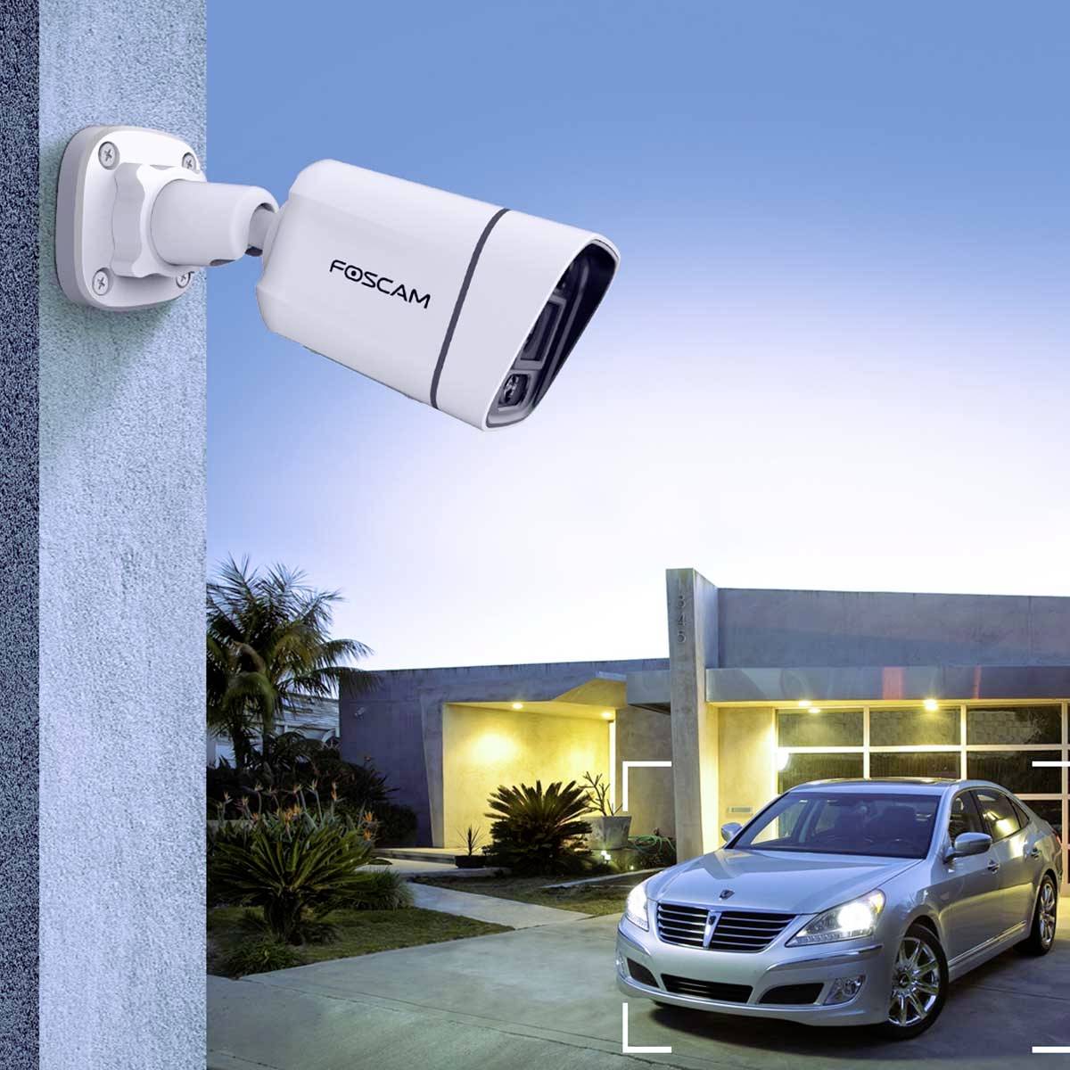 Security camera mounted on a house wall, monitoring driveway with parked car and illuminated building in the background.
