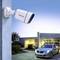 Security camera mounted on a house wall, monitoring driveway with parked car and illuminated building in the background.