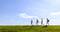 Four people are walking one behind the other across a green meadow under a clear, blue sky.