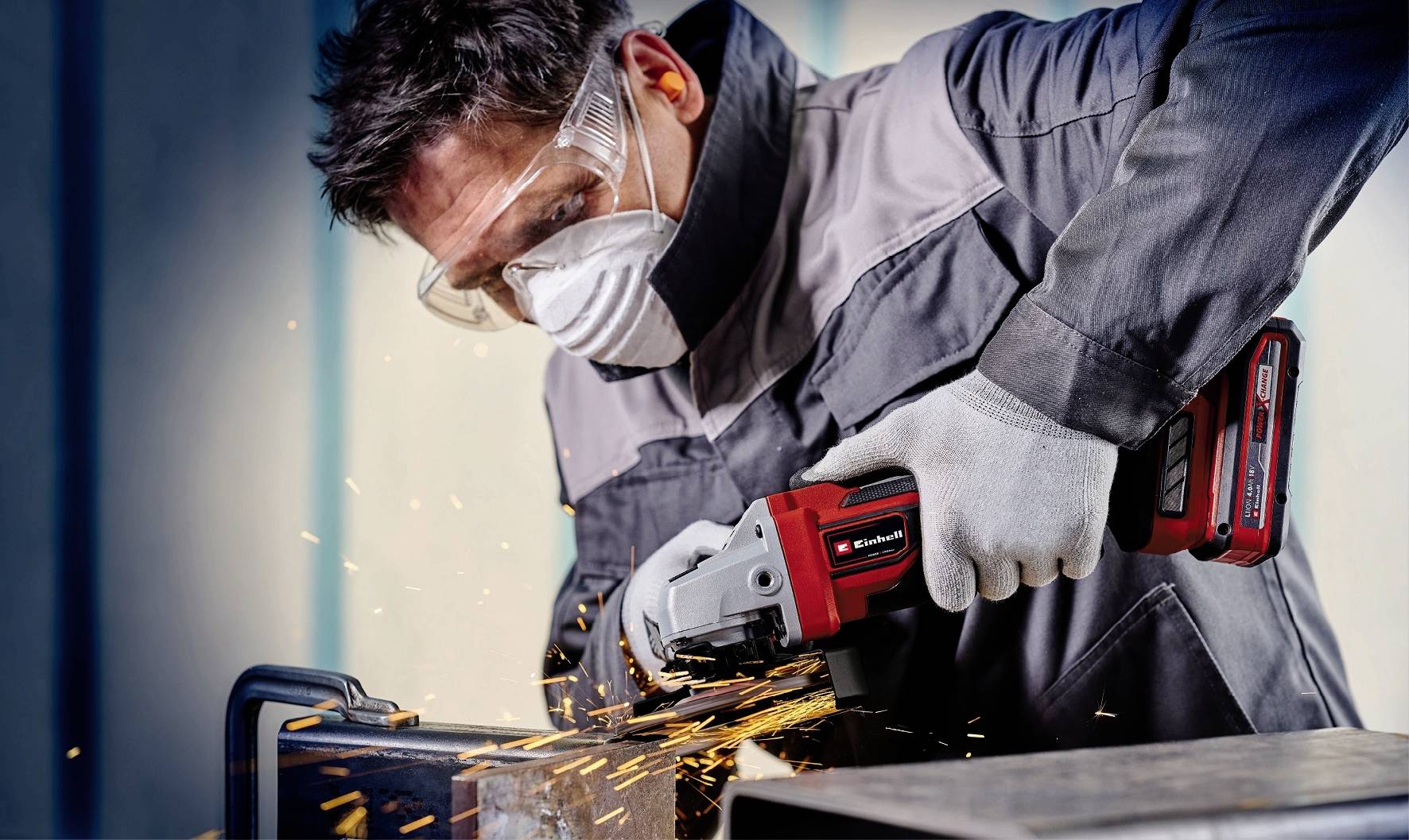 A tradesman is wearing protective clothing, safety glasses, and a mask while grinding sparks from a metal piece with an angle grinder.