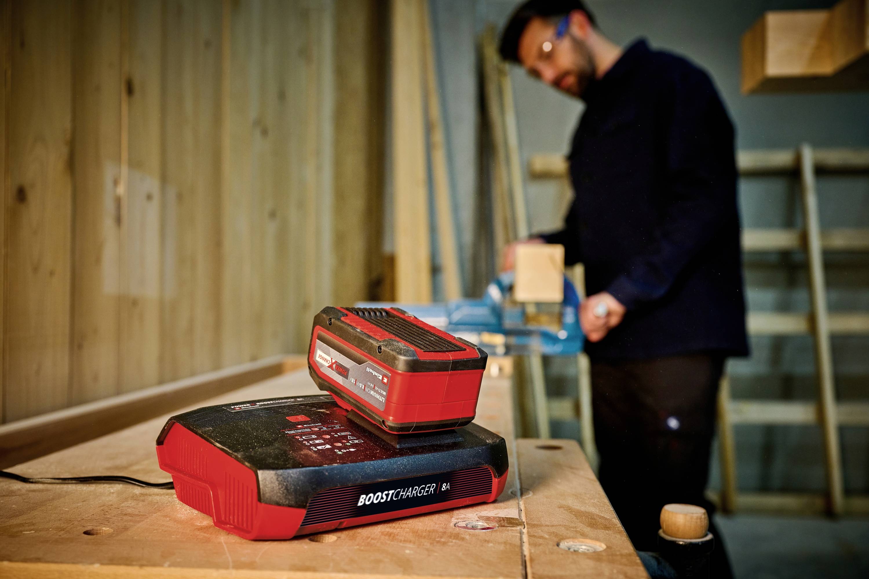 A red charger with a battery attached sits in the foreground on a table, while in the background a person is working with wood.
