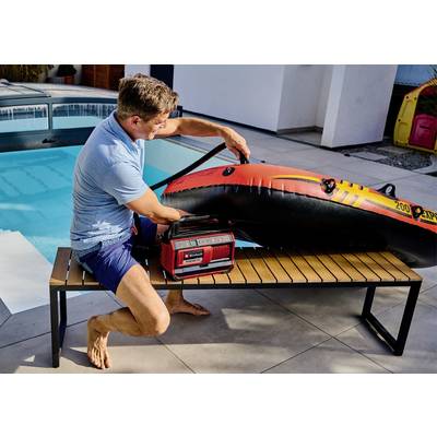 A man is inflating a large, orange inflatable dinghy with a portable air pump while sitting on a bench beside a swimming pool.