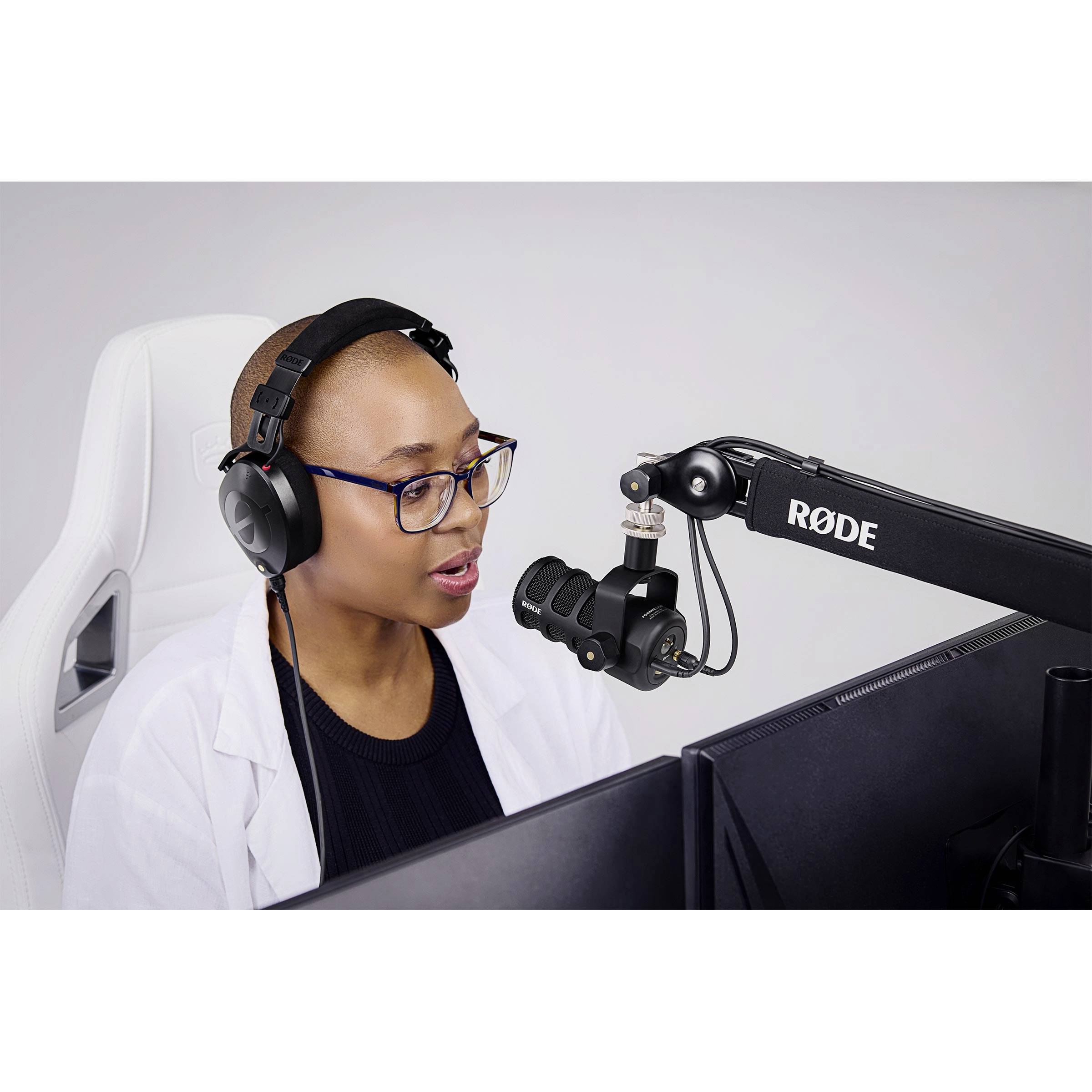 A person wearing headphones sits in front of a microphone and screen, speaking. They appear to be working in a recording studio or similar space.