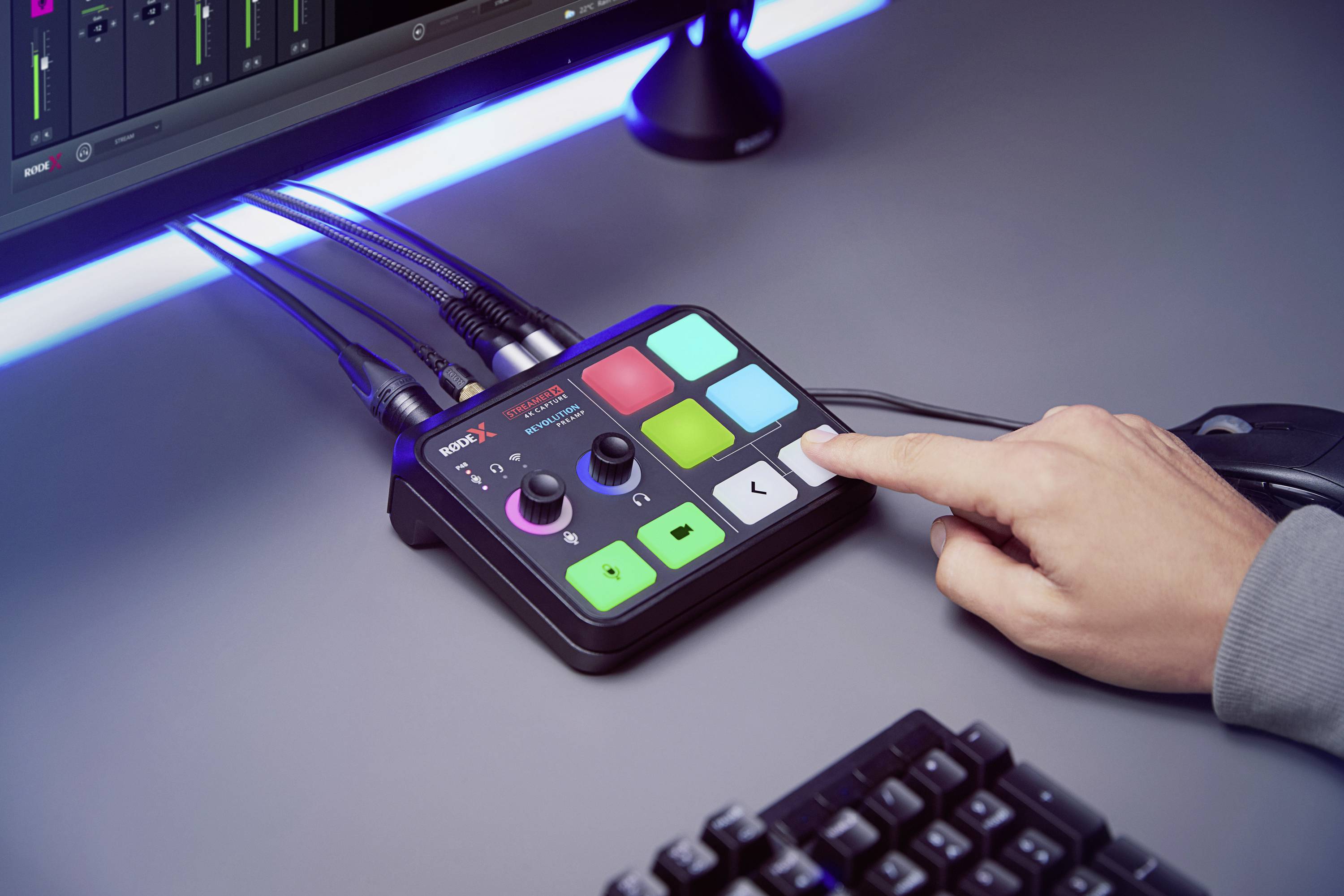 A person presses a button on an audio mixing desk next to a computer screen. Bright, colourful buttons light up on the device.