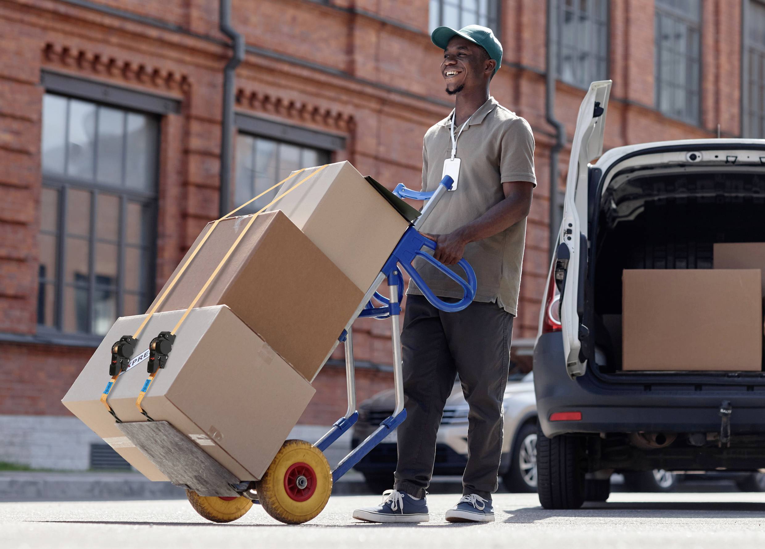 A man is transporting several boxes on a trolley in front of a brick building, smiling. In the background, an open delivery vehicle is parked.