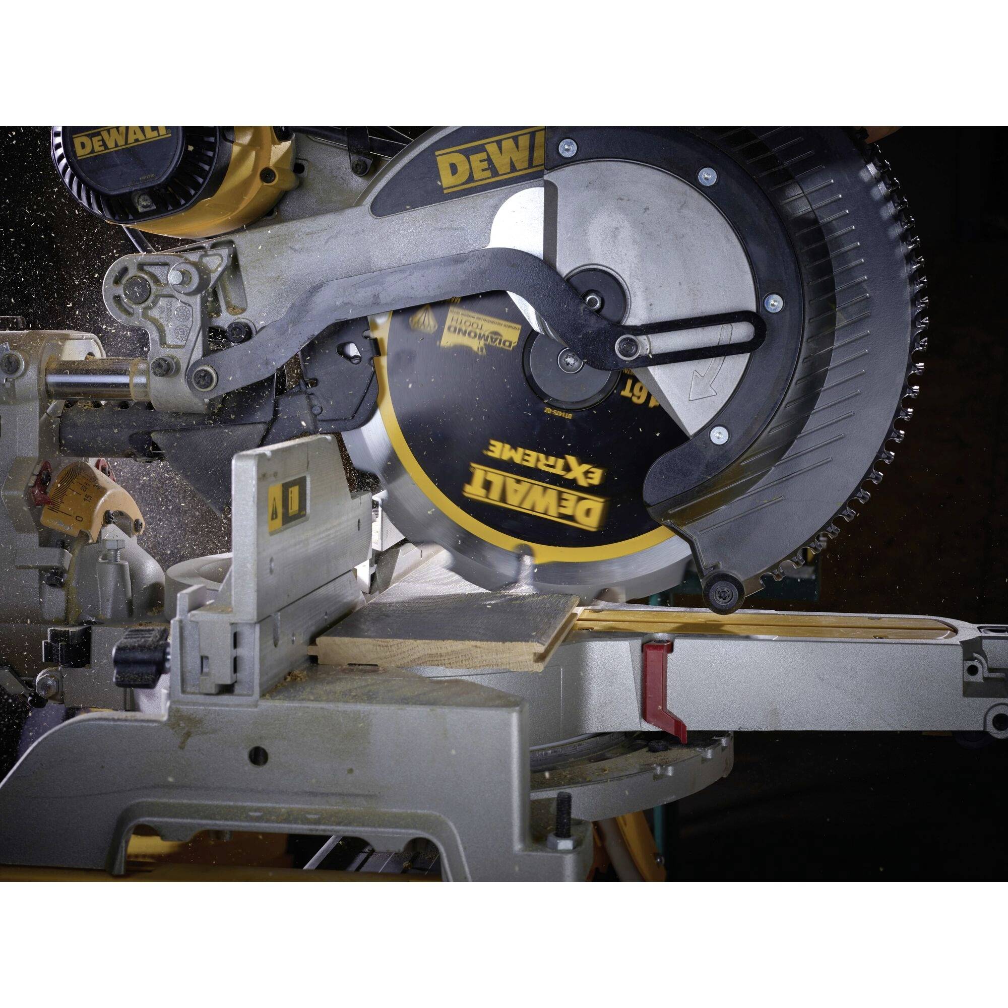 An electric mitre saw is cutting a wooden board. Wood shavings are flying through the air. The saw has a yellow casing and black details.